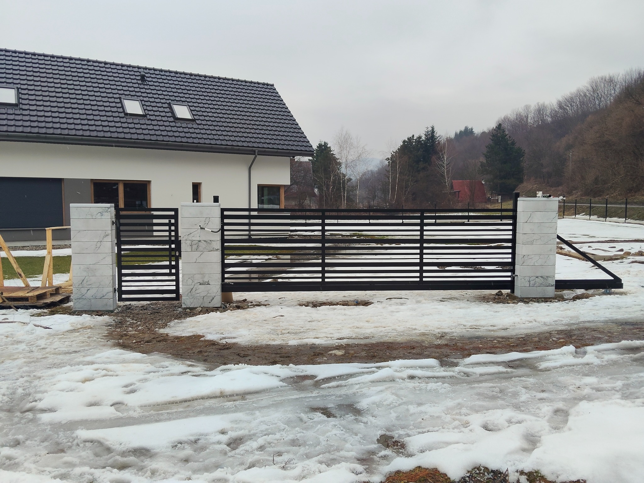 Nowoczesna, pozioma brama wjazdowa z czarnych profili, osadzona na słupkach z białego kamienia, na tle domu z ciemną dachówką i zimowego krajobrazu.