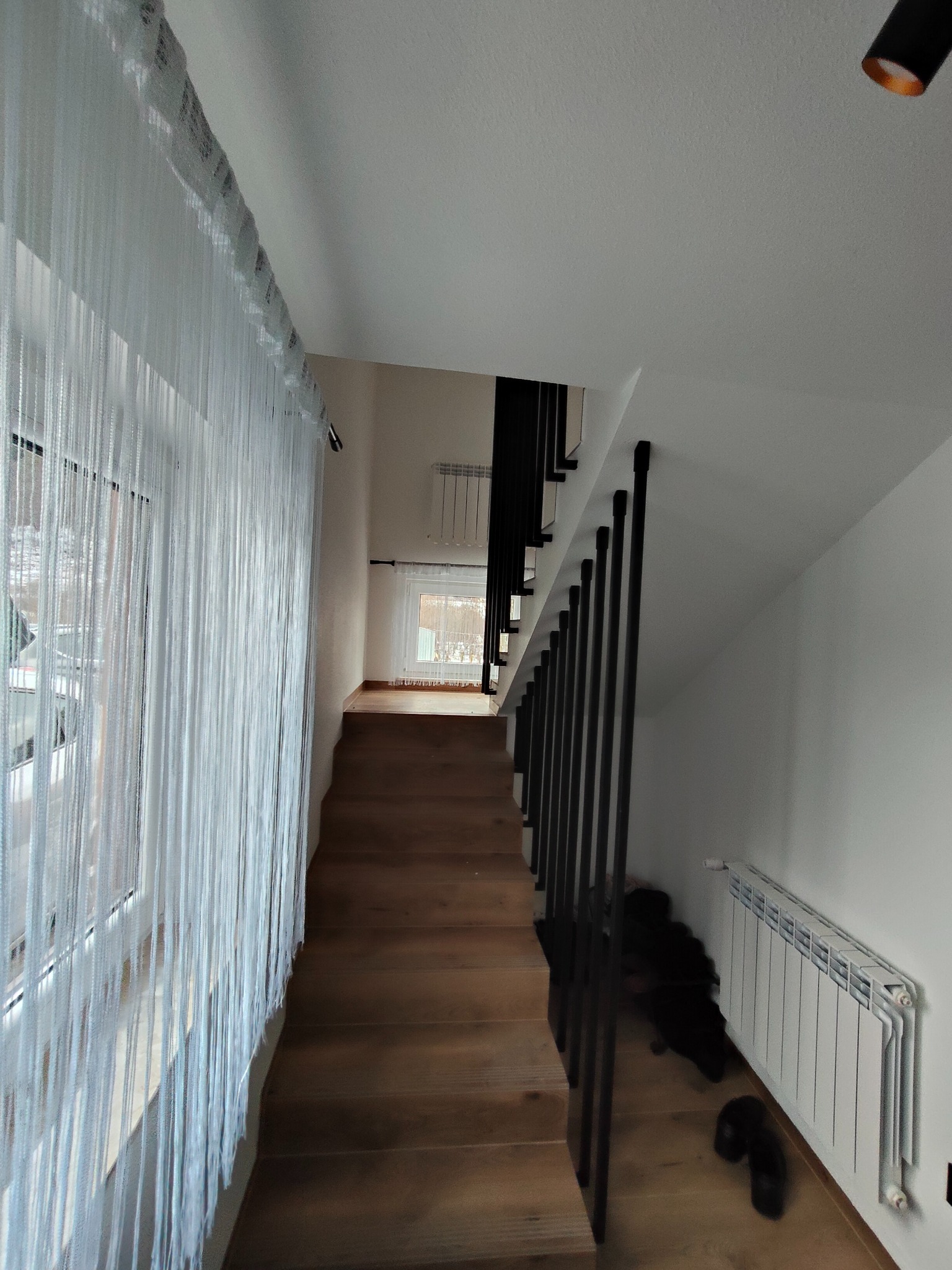 Nowoczesne schody drewniane z czarną, minimalistyczną balustradą metalową. Widok z góry, z dekoracyjną zasłoną sznurkową przy oknie. Jasne wnętrze.