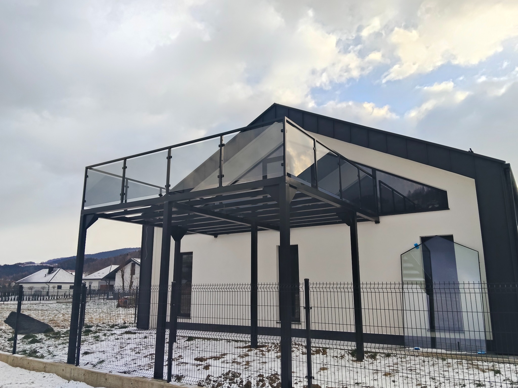 Nowoczesny dom z czarną metalową konstrukcją balkonu i szklanymi balustradami, otoczony metalowym ogrodzeniem panelowym na tle zimowego krajobrazu.
