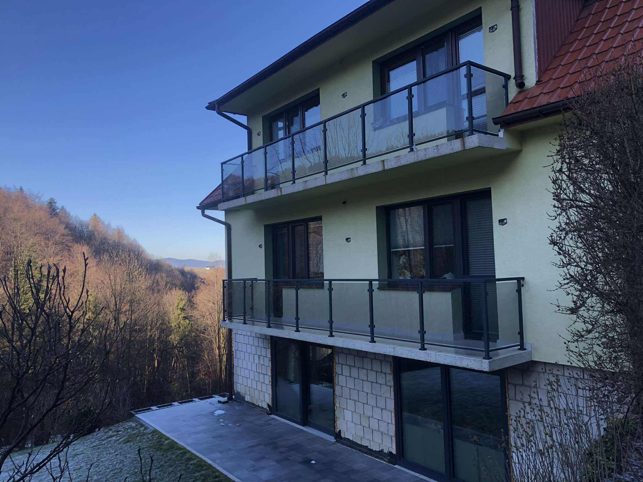 Dwukondygnacyjny dom z balkonami z balustradami szklanymi na tle zalesionego wzgórza. Balustrady z czarnymi ramami. Dolna kondygnacja w trakcie wykończenia.