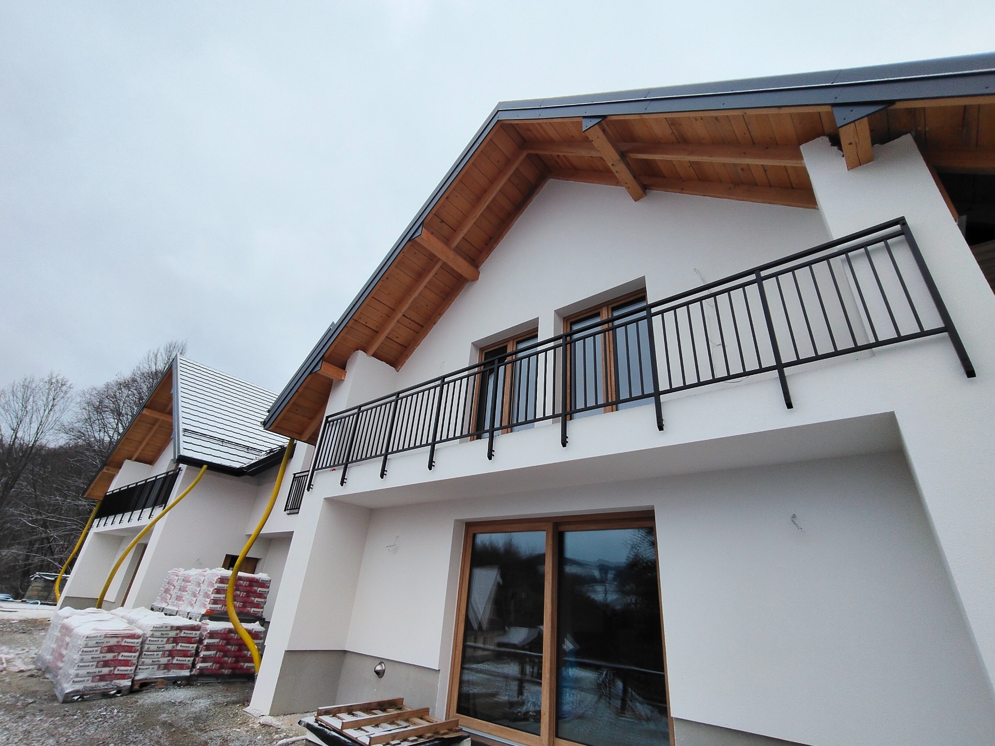 Nowoczesny balkon z czarną, metalową barierką w nowym budynku z drewnianym wykończeniem dachu. Na dole widoczne okno tarasowe i materiały budowlane.