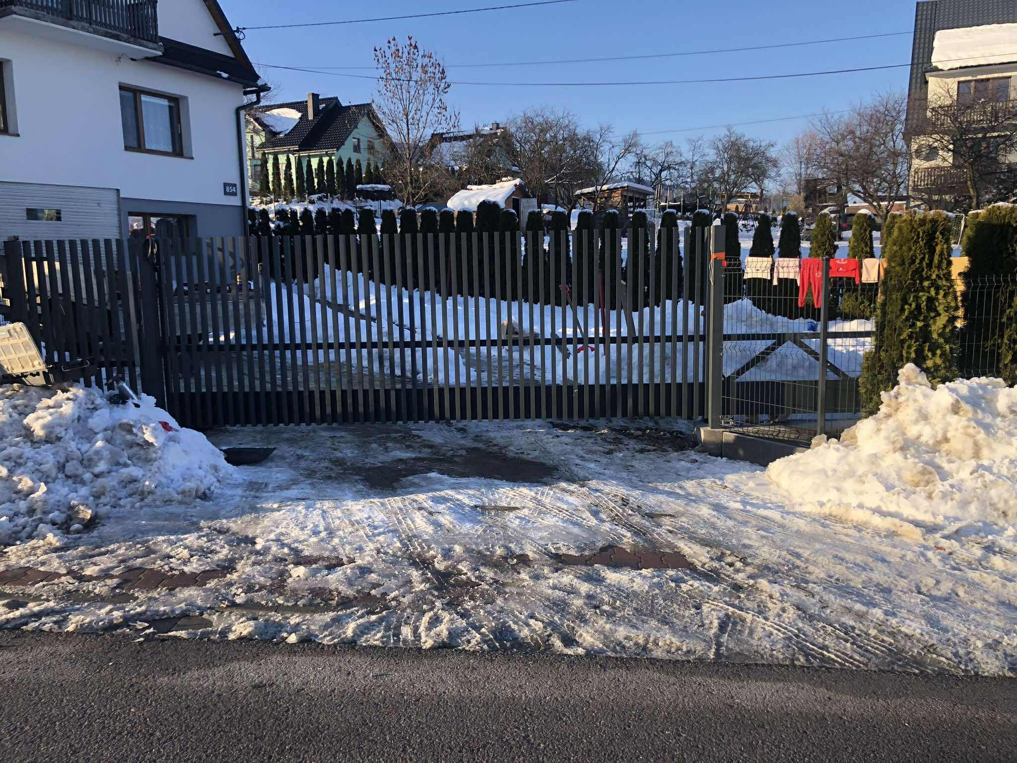 Nowoczesna, grafitowa brama wjazdowa z pionowych profili, z widocznymi śladami opon na śniegu i topniejącym śniegiem po bokach, na tle domów i zielonych tuj.