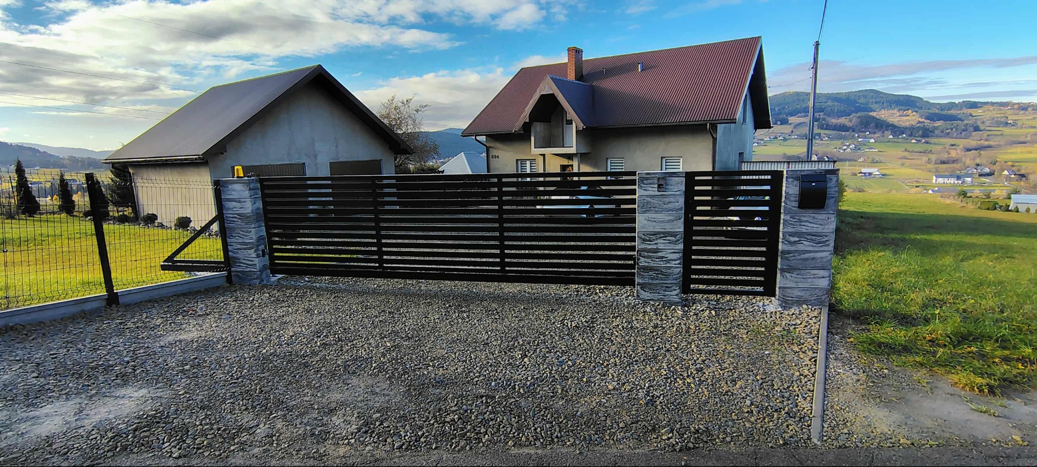 Nowoczesna brama wjazdowa z poziomych profili, osadzona na kamiennych słupkach z wbudowaną skrzynką na listy. Tło stanowi dom i malowniczy krajobraz.