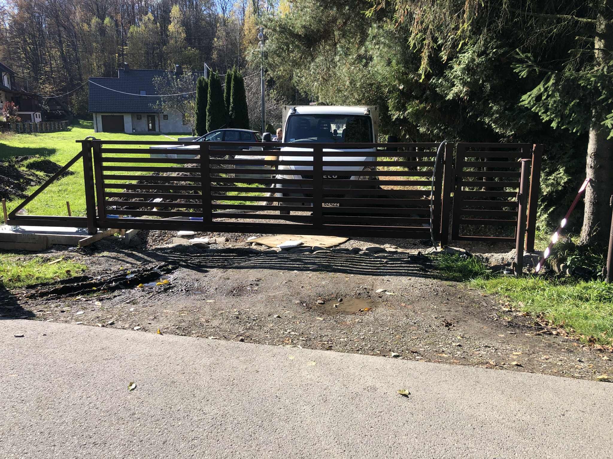 Nowoczesna brama wjazdowa z poziomych profili, częściowo zamontowana. Widoczny samochód dostawczy w tle i elementy montażowe. Ujęcie z poziomu ulicy.