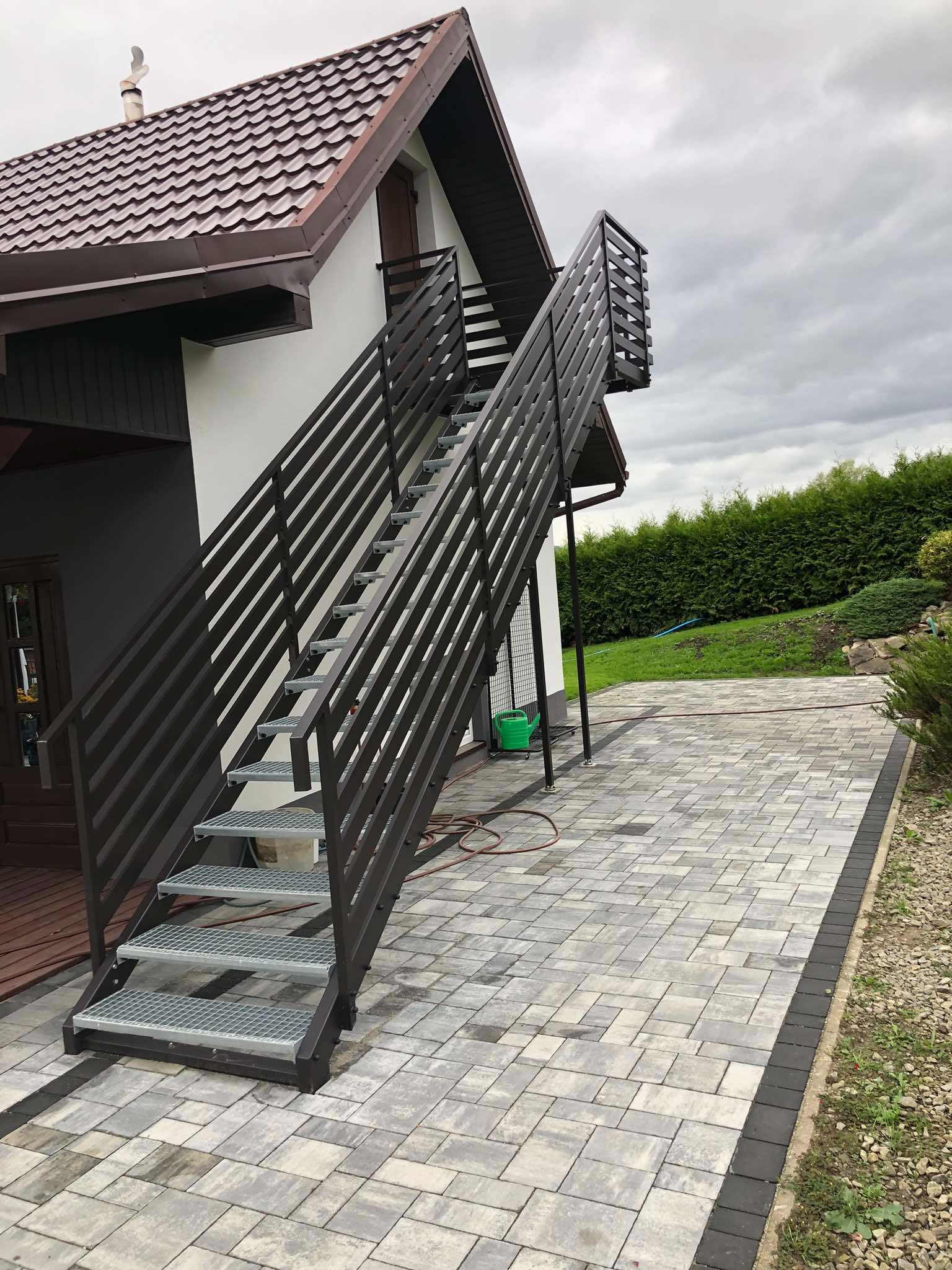 Zewnętrzne schody metalowe z brązową balustradą, prowadzące do drzwi na piętrze domu. Schody z ażurowymi stopniami, ułożone na kostce brukowej. Widok z dołu, pochmurne niebo.