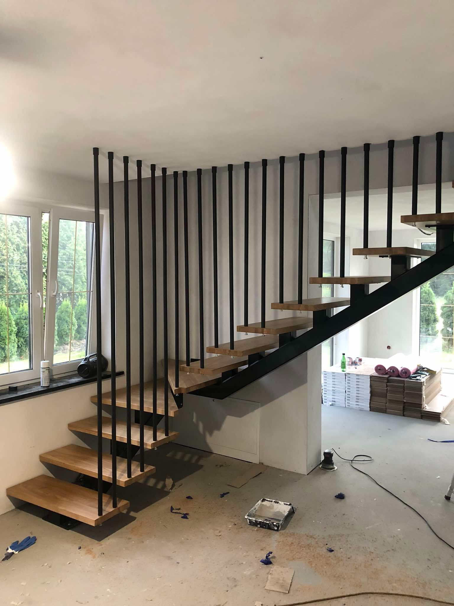 Nowoczesne schody metalowe z drewnianymi stopniami i czarną balustradą z pionowych prętów. Widok na okno i wnętrze domu. Podłoga w trakcie wykończenia.