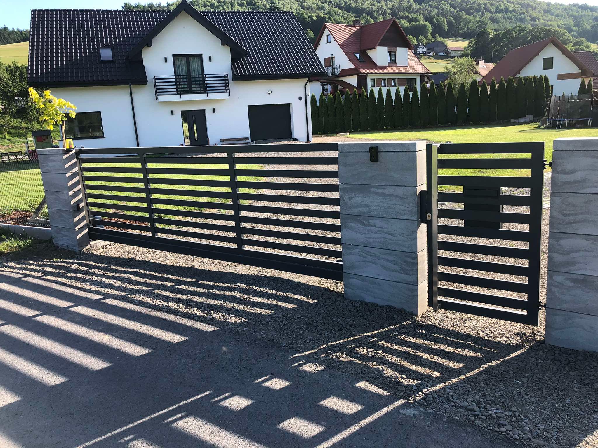 Nowoczesna, pozioma brama wjazdowa z szarego metalu, osadzona na kamiennych słupkach. W tle biały dom z czarnym dachem i zielone wzgórza.