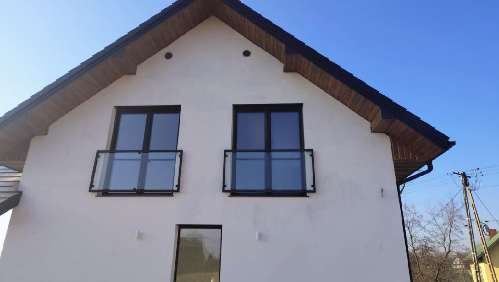 Biały dom z dwoma balkonami z czarnymi ramami i szklanymi balustradami. Ujęcie z dołu, podkreślające nowoczesny design i symetrię fasady.