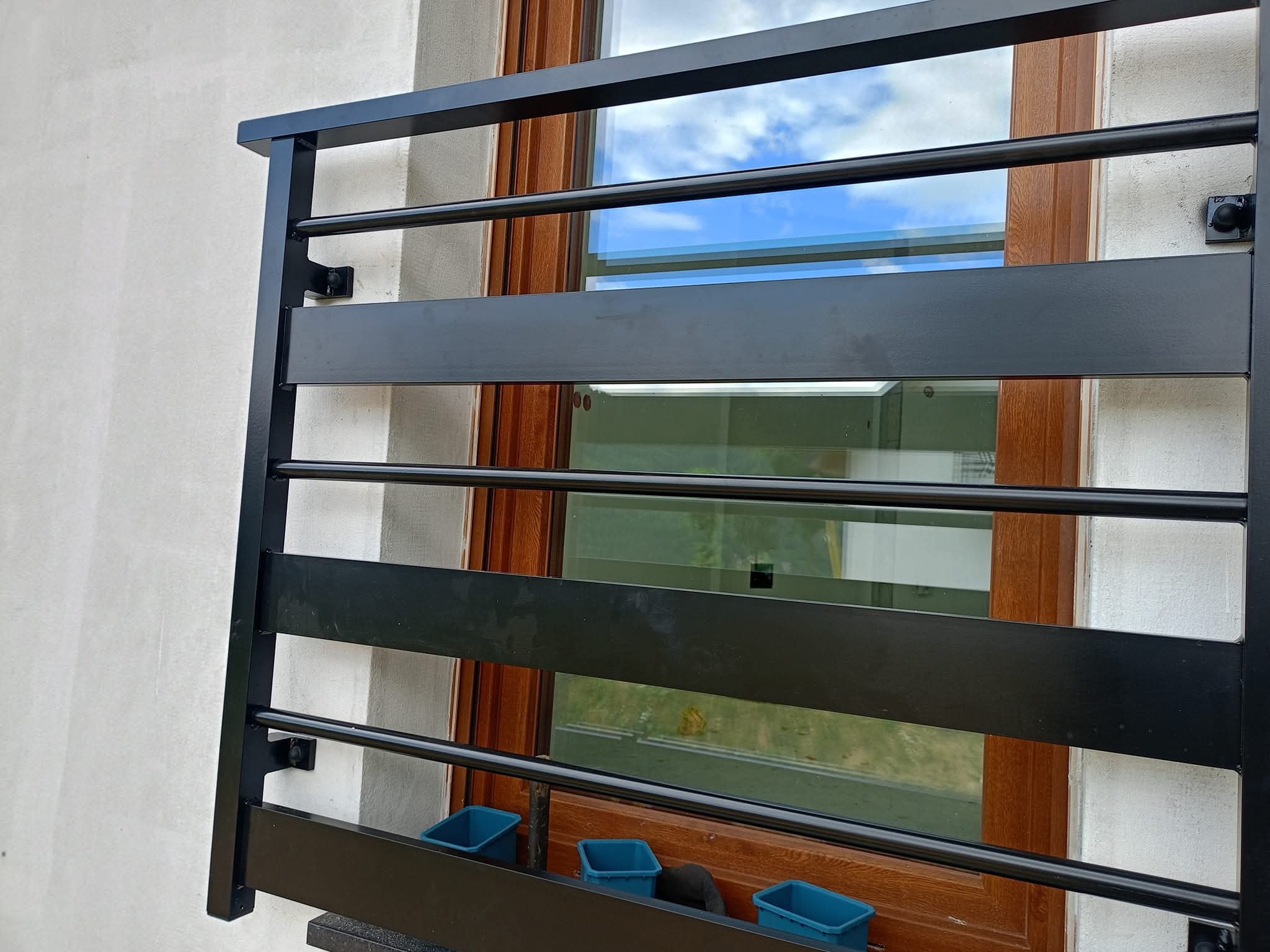 Czarna, metalowa balustrada balkonowa z poziomymi elementami, zamontowana przy oknie z drewnianą ramą. Widoczne niebieskie doniczki na kwiaty.