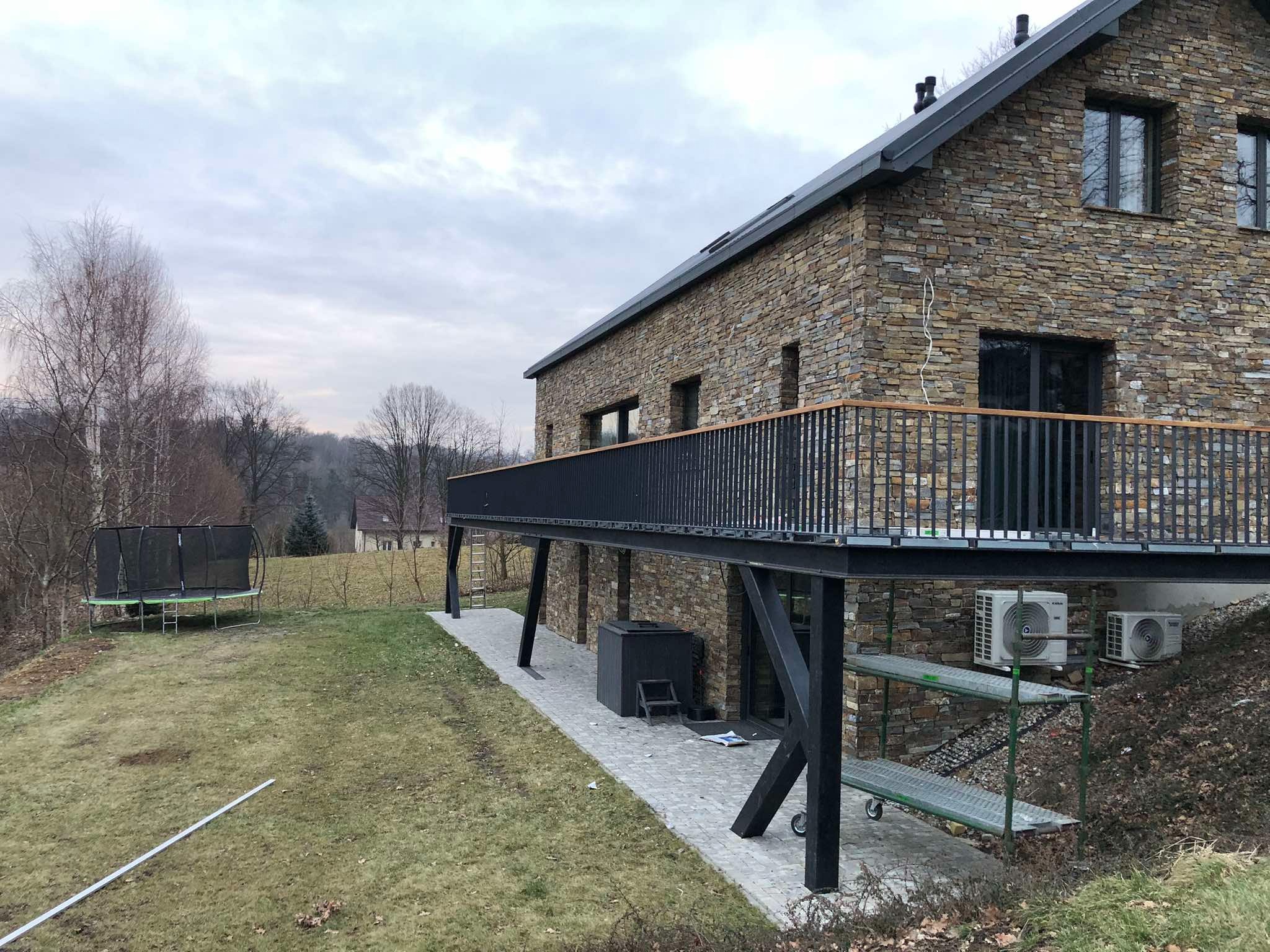 Nowoczesny balkon z czarną, metalową balustradą i drewnianym wykończeniem, wsparty na metalowych słupach, przylegający do kamiennego domu. Widok z zewnątrz.