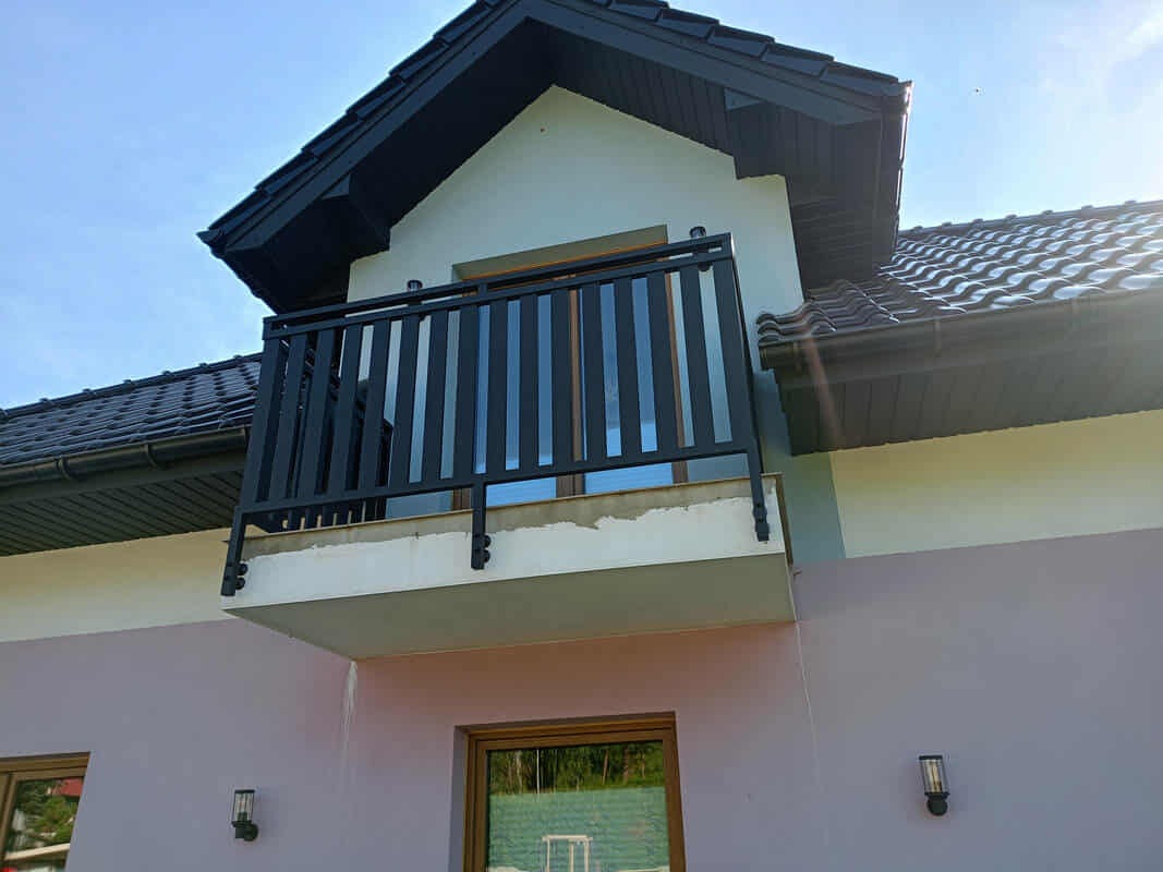 Czarna, metalowa balustrada balkonowa z pionowymi szczeblami, zamontowana na jasnym balkonie domu z ciemnym dachem, widziana z dołu pod kątem.