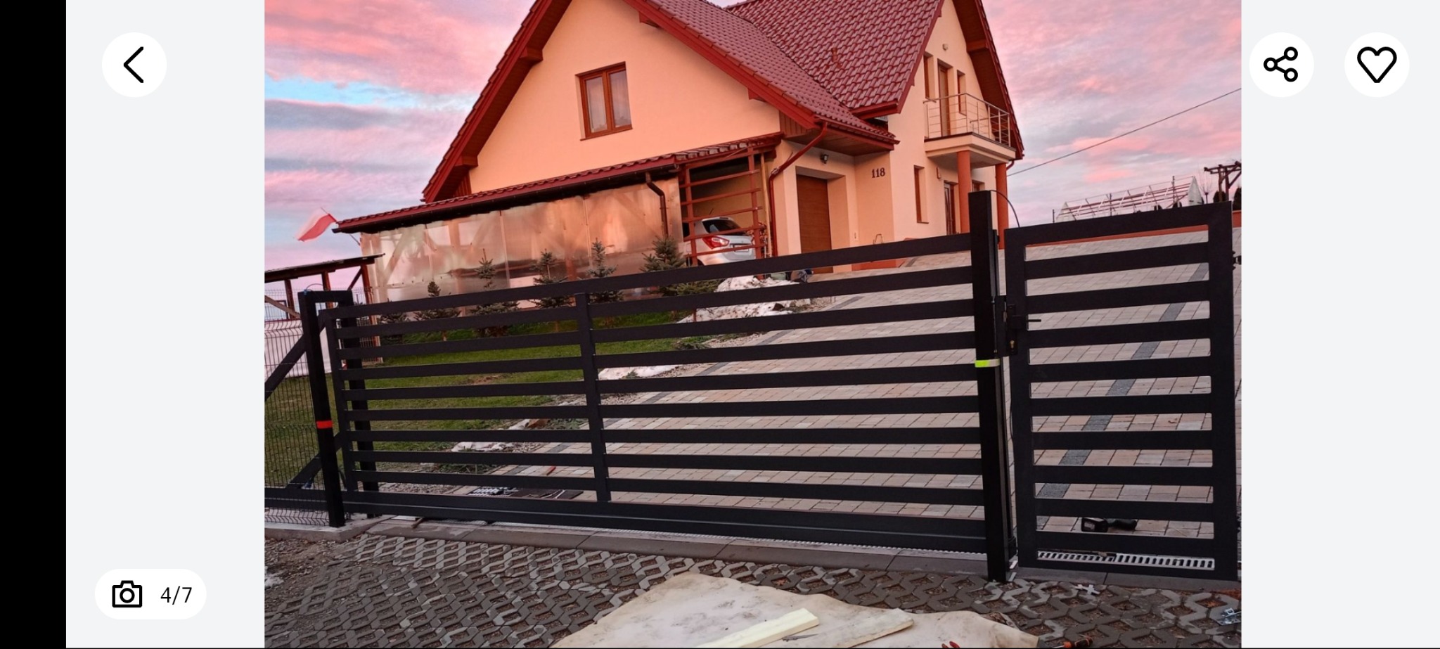 Nowoczesna, pozioma brama wjazdowa w kolorze antracytu, na tle domu z czerwoną dachówką i różowego nieba o zmierzchu. Widoczne detale konstrukcyjne i automatyka bramy.