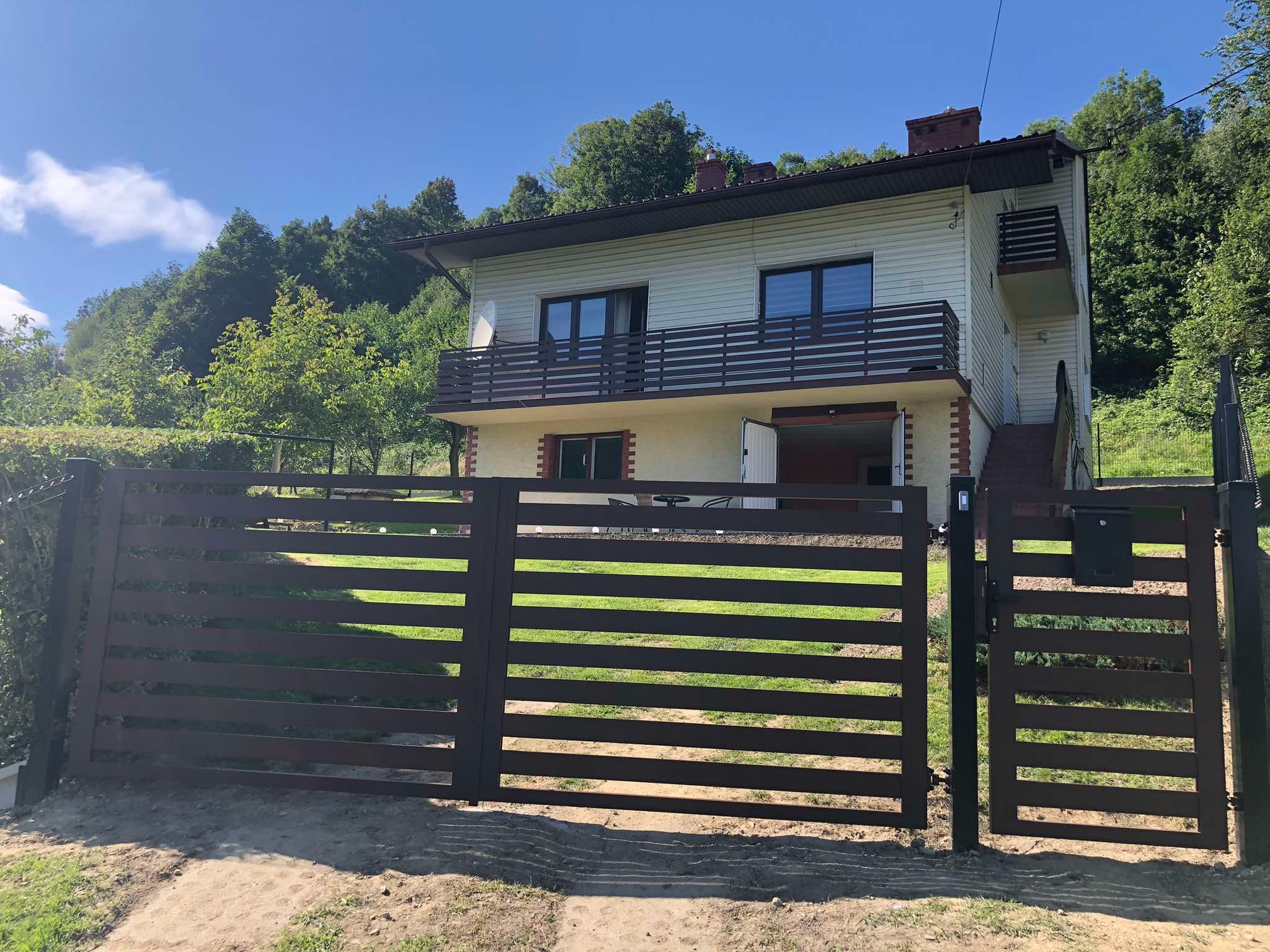 Nowoczesna, pozioma brama wjazdowa w kolorze ciemnego brązu, z furtką i skrzynką na listy, na tle domu z balkonem i zieleni. Słoneczny dzień.