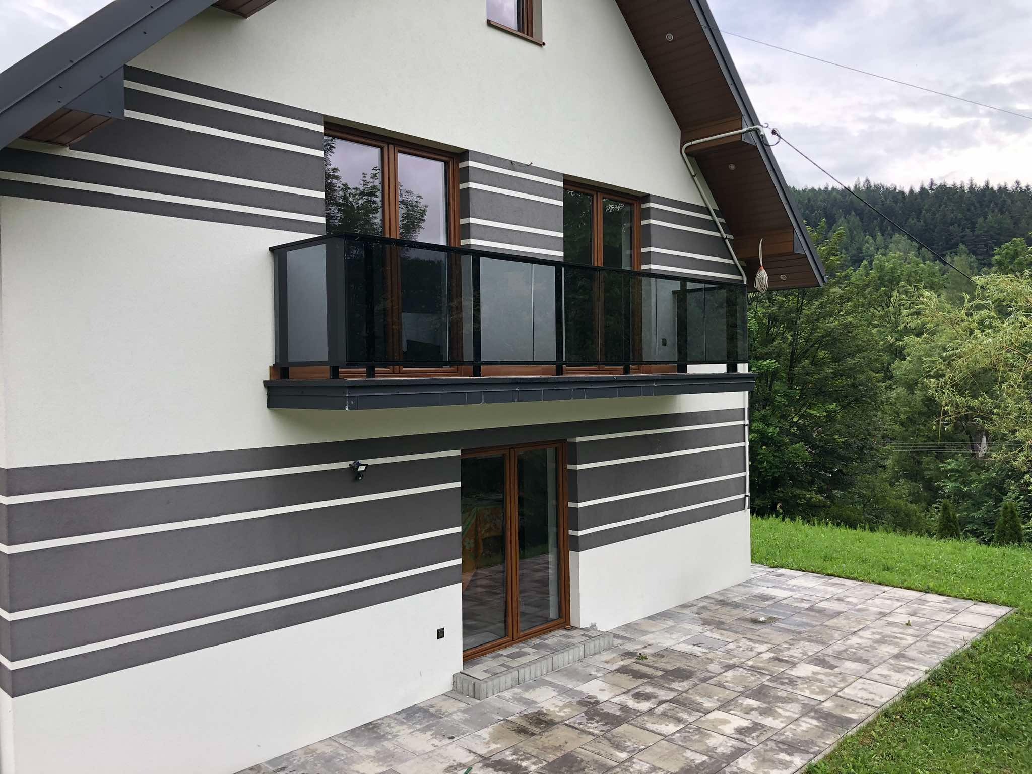 Nowoczesny balkon z balustradą szklaną i drewnianymi ramami, na tle domu z poziomymi pasami. Widok na zieleń i las w tle. Ujęcie z dołu, podkreślające architekturę.