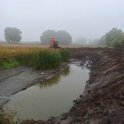 Wykopany staw w mglisty dzień, pomarańczowa koparka w tle, wysoka trawa i trzciny na pierwszym planie.
