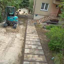 Ścieżka z kostki brukowej prowadząca do domu, z mini-koparką Sunward obok, teren przygotowany pod dalsze prace.