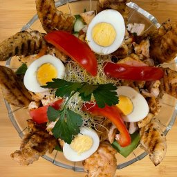 Planbar - Sałatka z grillowanym kurczakiem, jajkiem na twardo, papryką, ogórkiem i kiełkami, podana w szklanej misce na drewnianym stole.