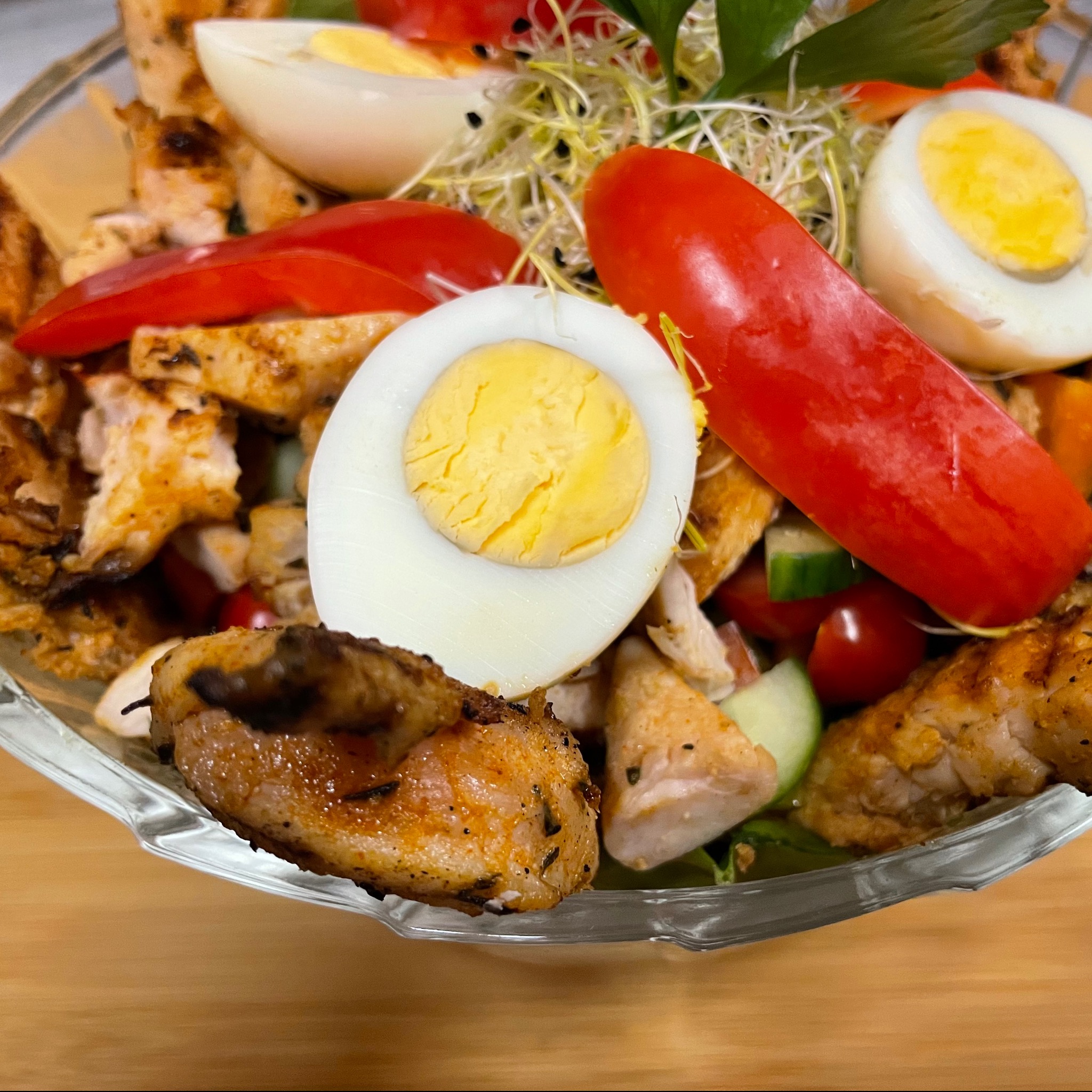 Szklana miska z sałatką: grillowany kurczak, jajko na twardo, czerwona papryka, ogórek, kiełki i pomidorki koktajlowe na drewnianym stole.
