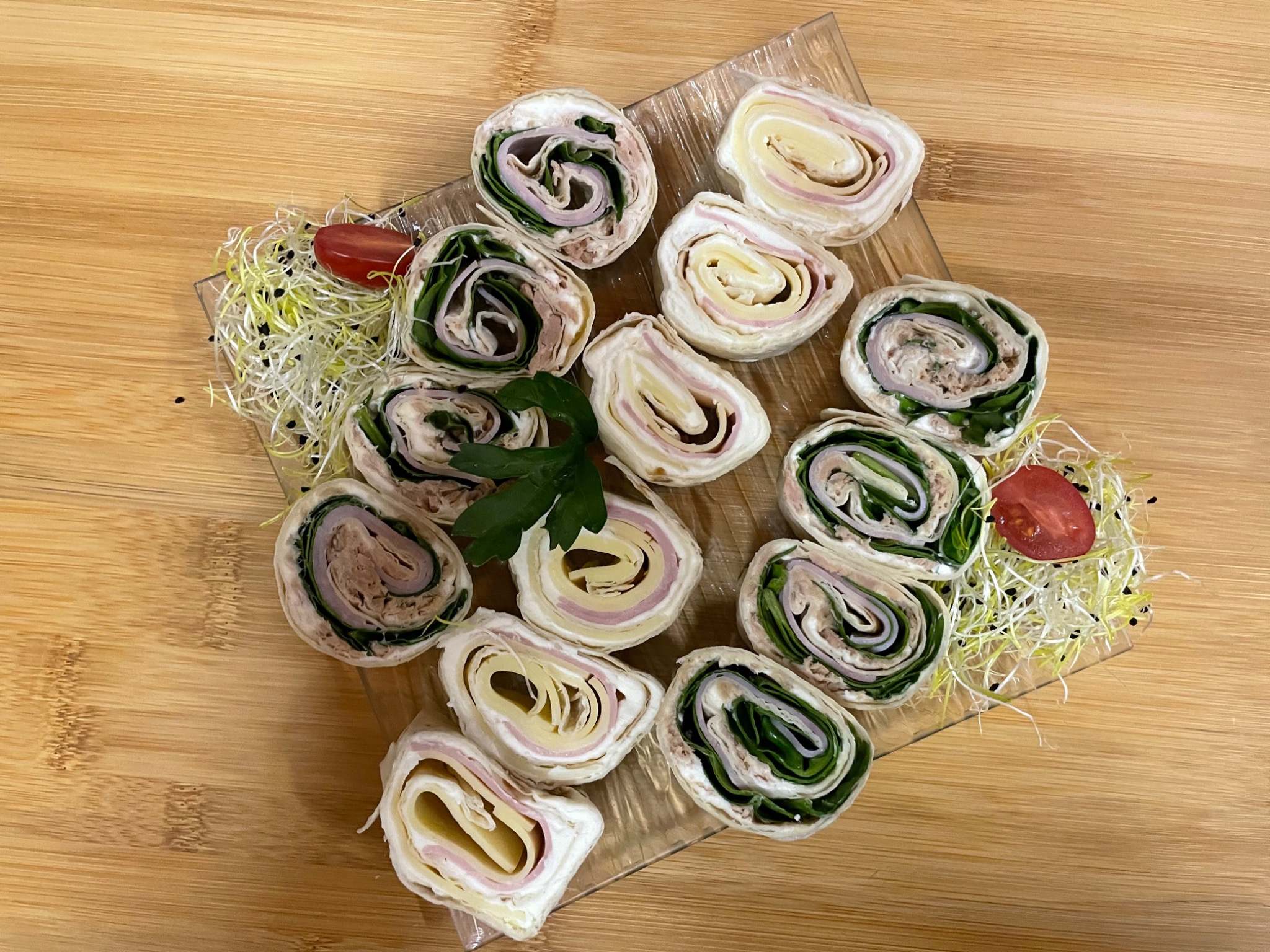 Dekoracyjnie ułożone roladki z tortilli z różnymi nadzieniami (szynka, ser, zielenina, pasta) na przezroczystym talerzu, ozdobione kiełkami i połówkami pomidorków koktajlowych, na drewnianym tle.