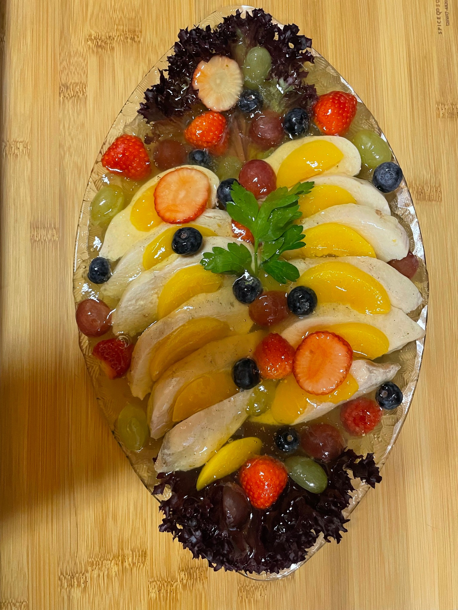 Deser owocowy w galarecie na szklanym półmisku, ozdobiony sałatą strzępiastą, truskawkami, winogronami, borówkami, brzoskwiniami i natką pietruszki, na drewnianym stole.