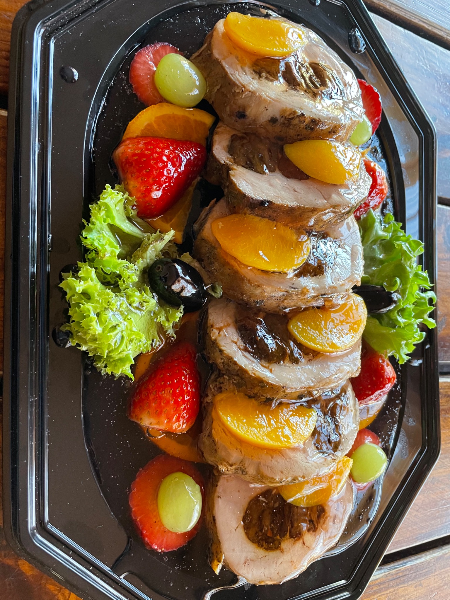 Rolada z jasnego mięsa w plastrach, udekorowana brzoskwiniami, truskawkami, winogronami, oliwkami i sałatą na czarnej, lakierowanej tacy z sosem.