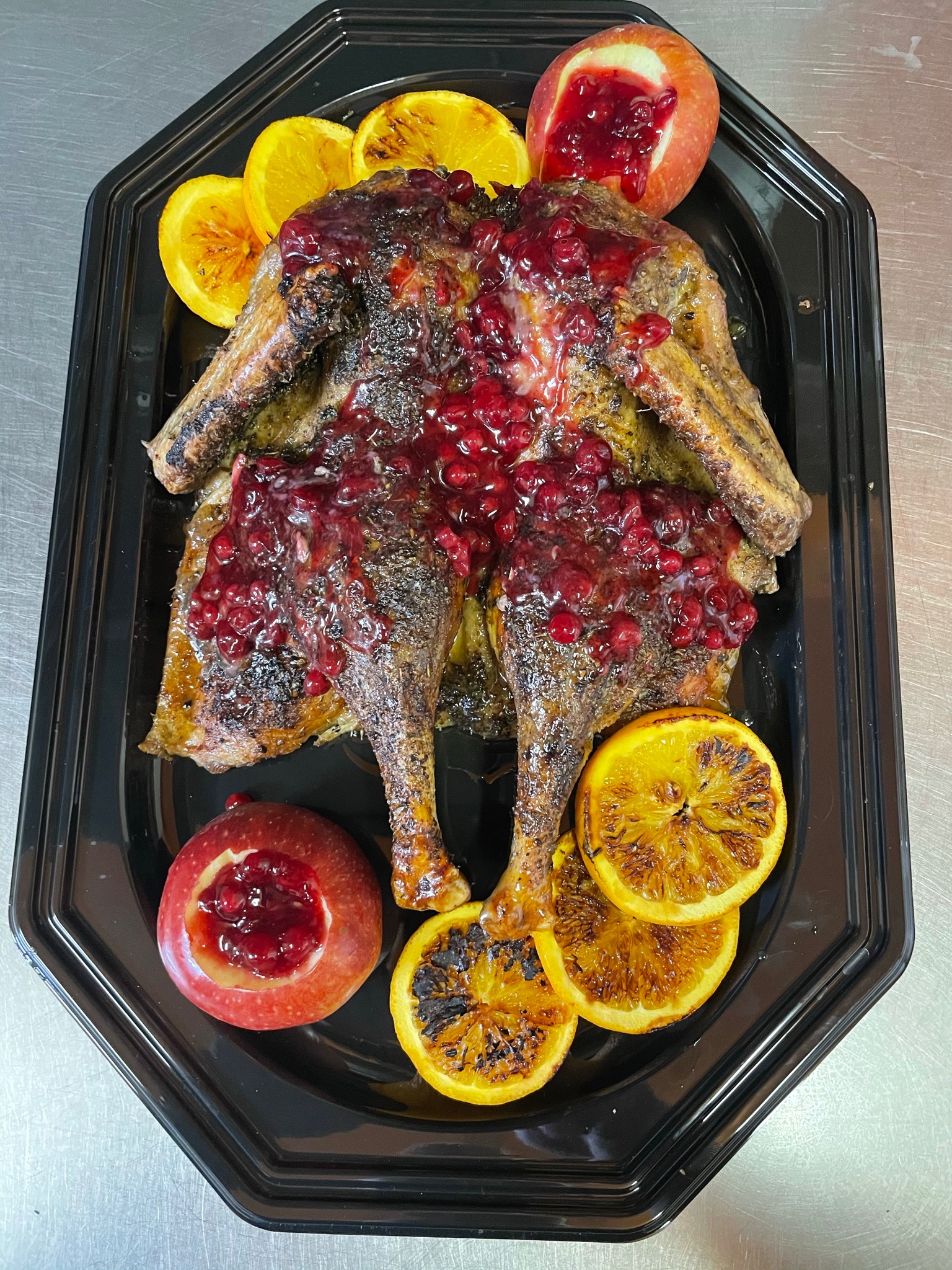 Pieczona kaczka polana sosem żurawinowym, dekorowana grillowanymi pomarańczami i jabłkami z żurawiną, ułożona na czarnej tacy.