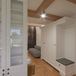 Adrianna - Wnętrze przedpokoju z białą szafą wnękową z siedziskiem, drewnianą podłogą ułożoną w jodełkę, widocznymi belkami stropowymi i przeszkloną szafą na lewo, oraz tapetą z motywem leśnym w tle.