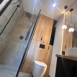 Ciężkowski Adam - Nowoczesna łazienka z kabiną prysznicową typu walk-in z czarnymi profilami, podłogą imitującą drewno, wiszącą toaletą i szafkami z drewnianym fornirem oraz czarnymi akcentami.