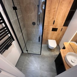 Ciężkowski Adam - Nowoczesna łazienka w Gdyni po remoncie, z kabiną prysznicową typu walk-in z czarnymi ramami, drewnianymi szafkami i umywalkami nablatowymi o organicznych kształtach.