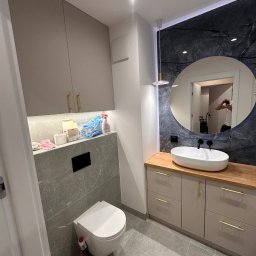 Ciężkowski Adam - Nowoczesna łazienka z podwieszaną toaletą, umywalką nablatową na drewnianym blacie i okrągłym lustrem na tle ściany z imitacją marmuru, oświetlona wiszącą lampą.