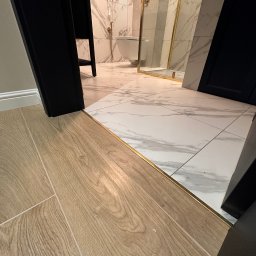 Ciężkowski Adam - Połączenie podłogi drewnianej z marmurową w wyremontowanej łazience z kabiną prysznicową ze złotymi elementami, widoczne przejście z listwą wykończeniową.