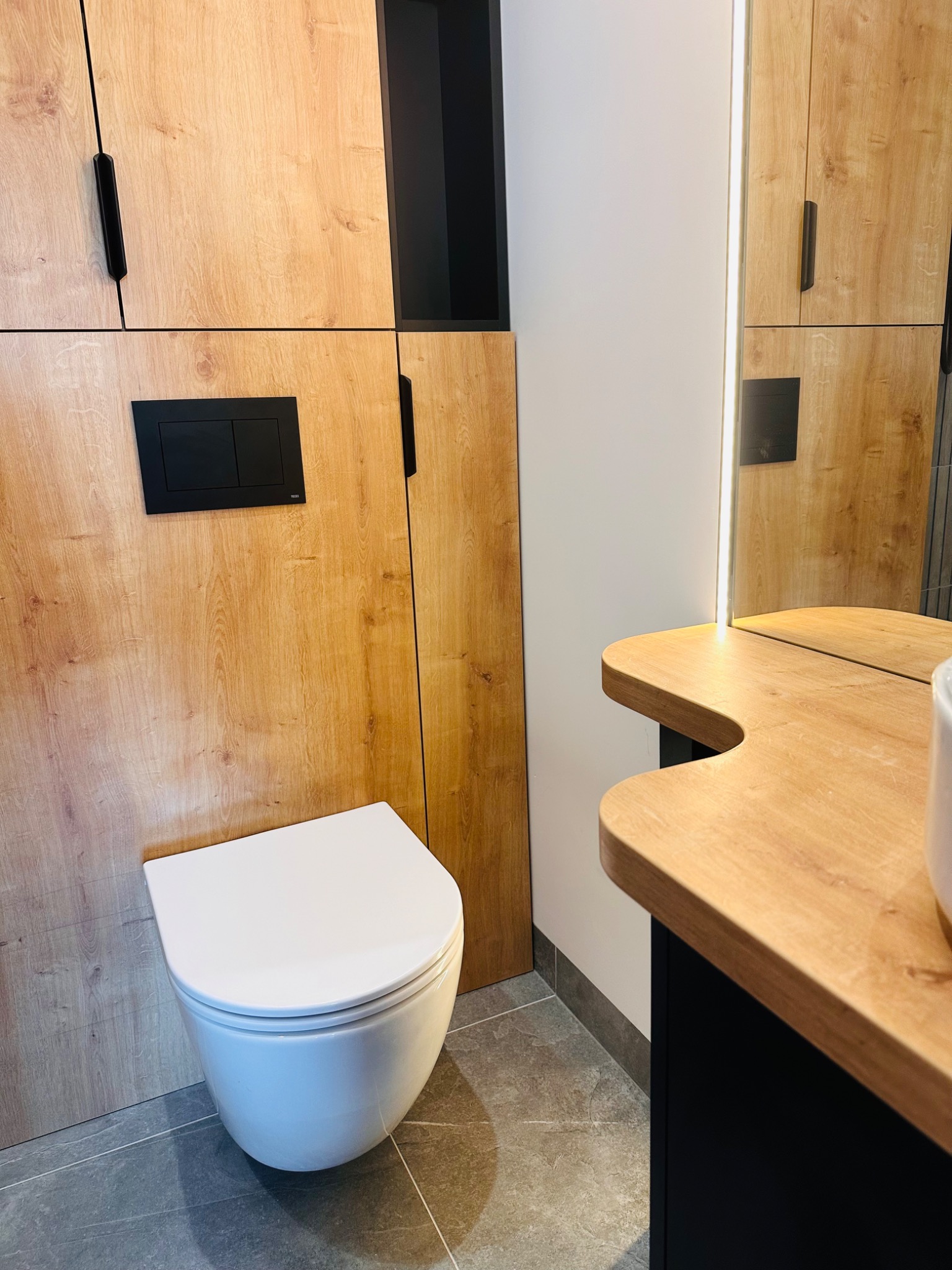 Nowoczesna łazienka z podwieszaną miską WC, szafką z drewnopodobnym frontem i czarnymi akcentami, oświetlenie LED przy lustrze.