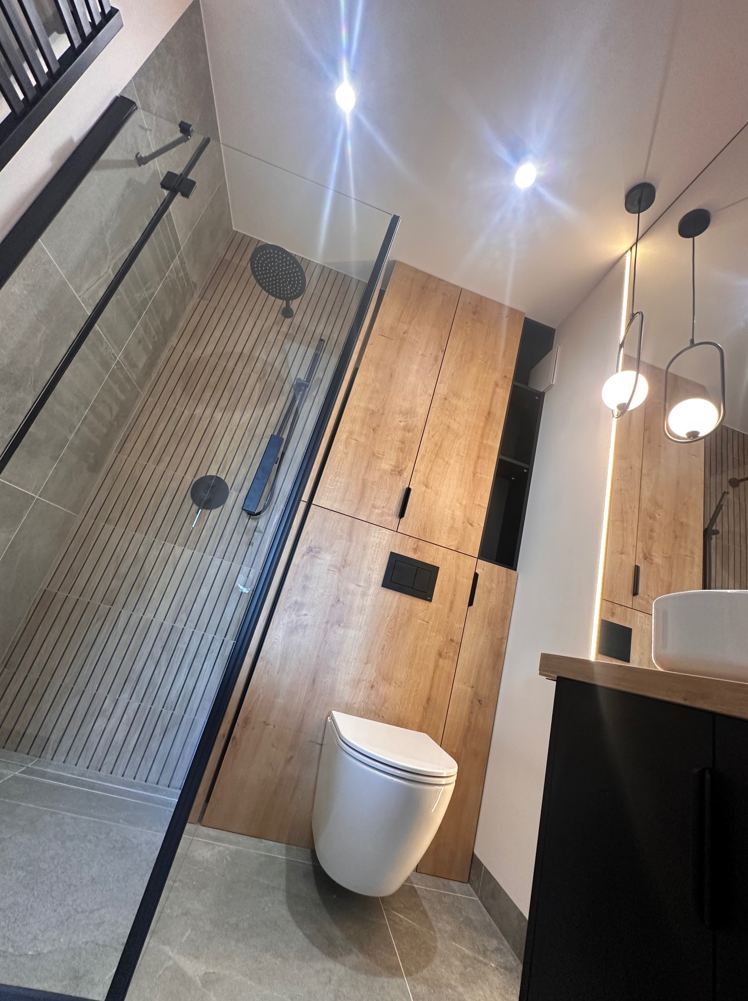 Nowoczesna łazienka z kabiną prysznicową typu walk-in z czarnymi profilami, podłogą imitującą drewno, wiszącą toaletą i szafkami z drewnianym fornirem oraz czarnymi akcentami.