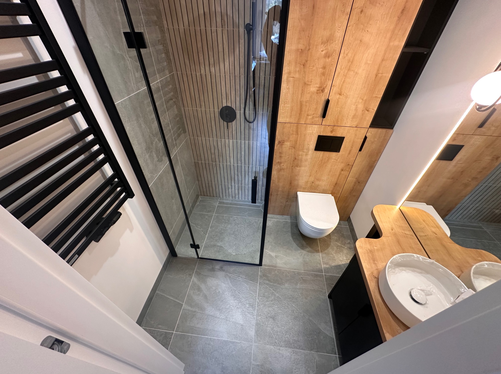 Nowoczesna łazienka w Gdyni po remoncie, z kabiną prysznicową typu walk-in z czarnymi ramami, drewnianymi szafkami i umywalkami nablatowymi o organicznych kształtach.