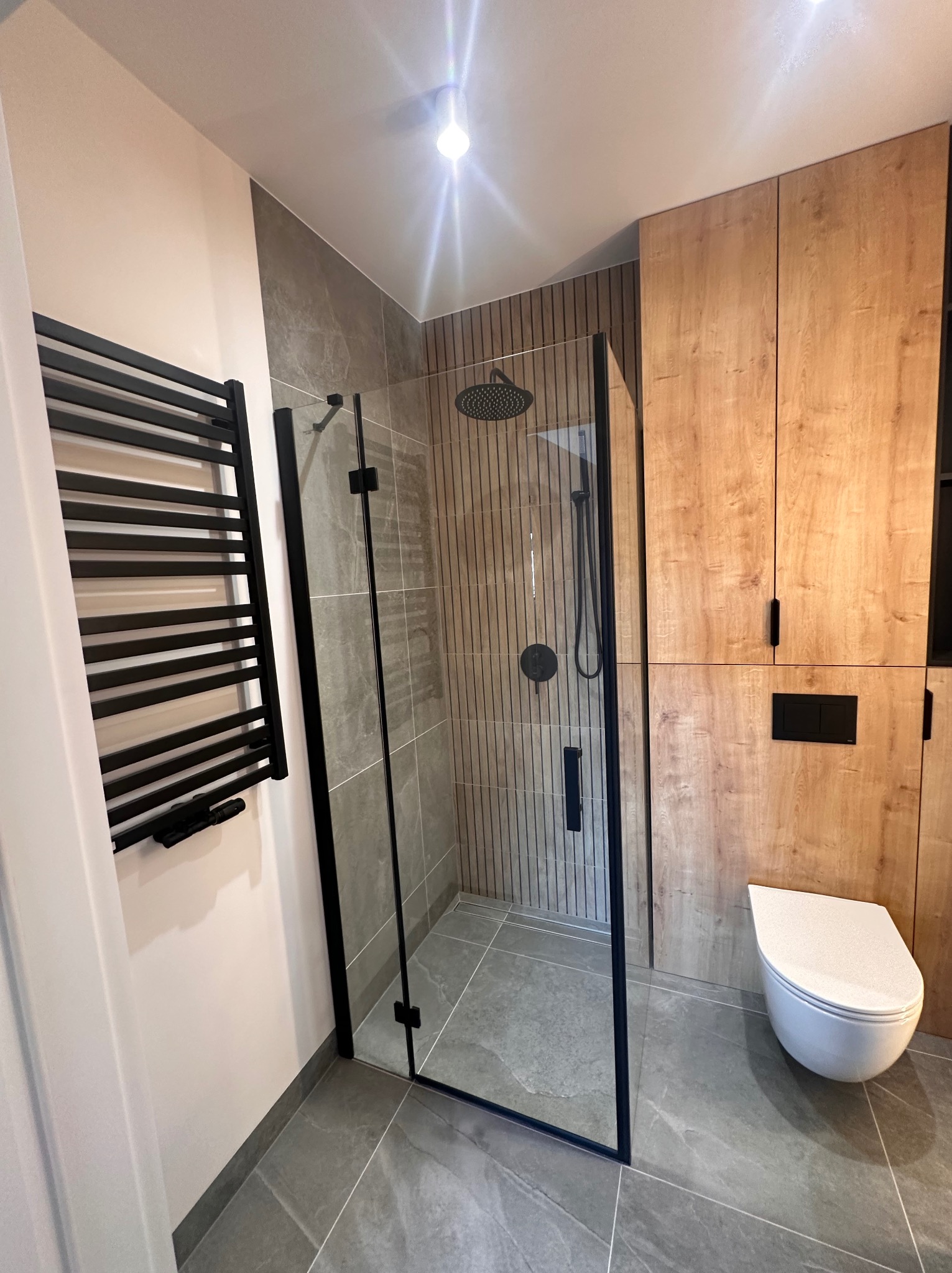 Nowoczesna łazienka z kabiną prysznicową typu walk-in z czarnymi profilami, grzejnikiem drabinkowym w czarnym kolorze, drewnianymi szafkami i szarą podłogą z płytek imitujących kamień.