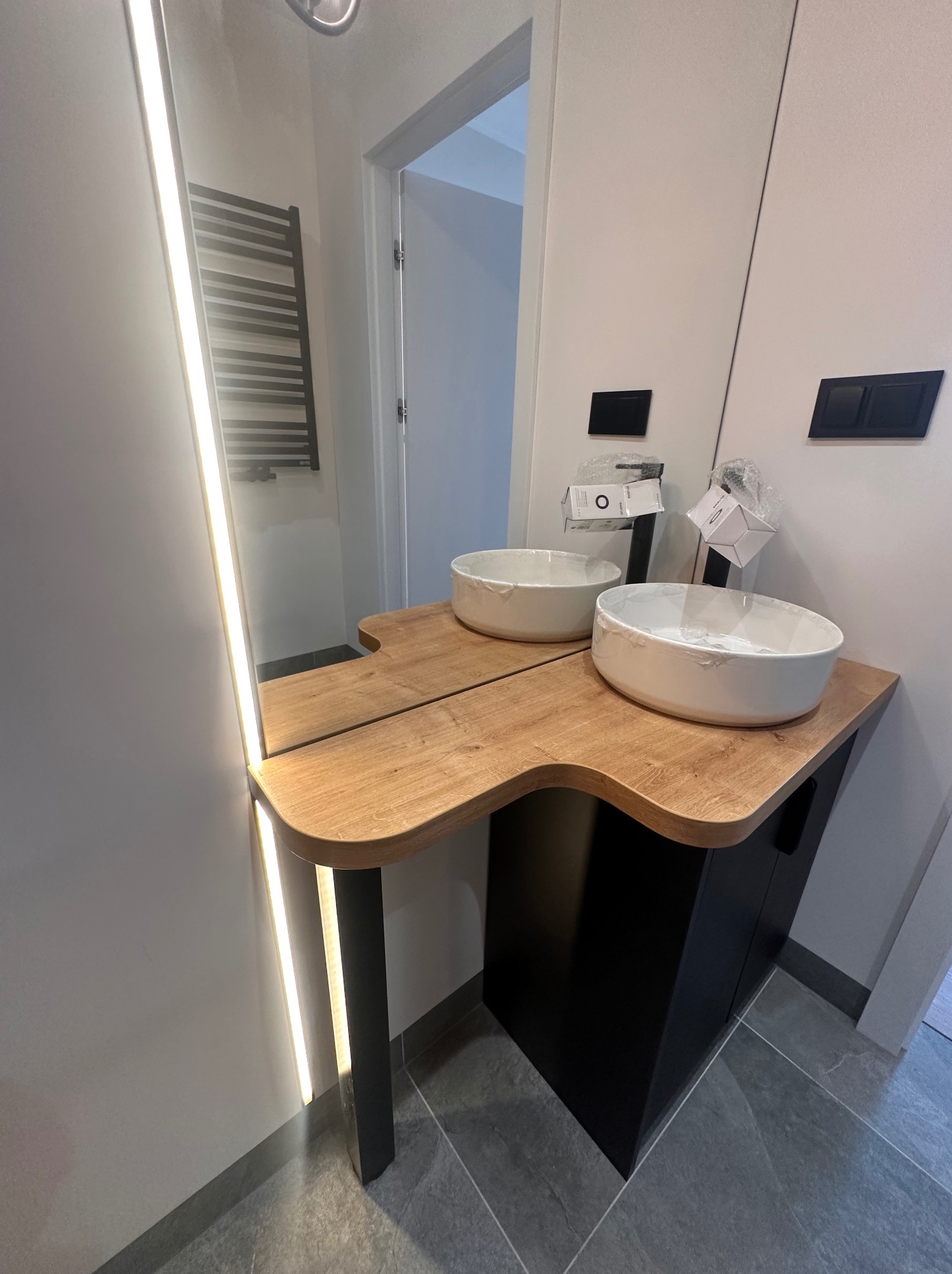 Nowoczesna łazienka z dwiema owalnymi umywalkami nablatowymi na drewnianym blacie z czarną szafką, oświetlenie LED w lustrze i pod blatem, widoczny grzejnik drabinkowy na ścianie.