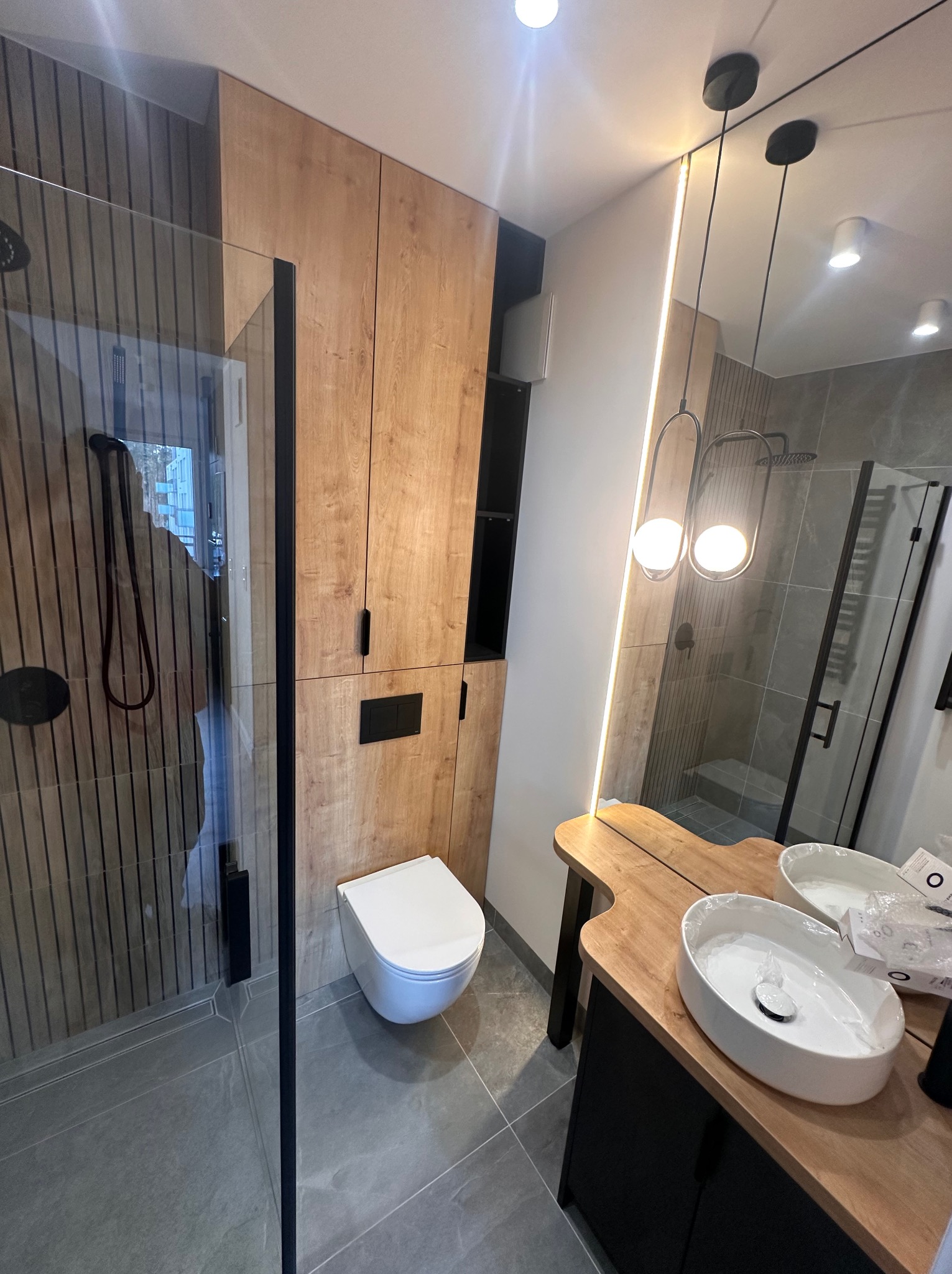 Nowoczesna łazienka z kabiną prysznicową typu walk-in, podwieszaną toaletą i drewnianą zabudową meblową, oświetlona lampami wiszącymi z kulistymi kloszami.