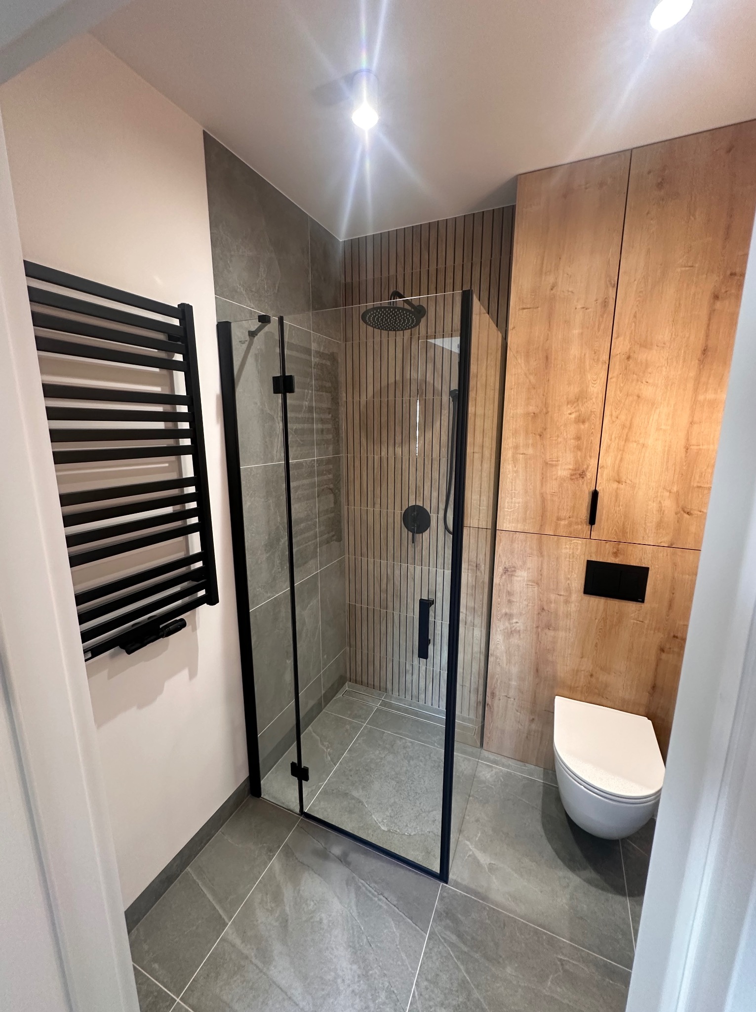Nowoczesna łazienka z kabiną prysznicową typu walk-in z czarnymi profilami, grzejnikiem drabinkowym w kolorze czarnym, szarą podłogą i ścianami wykończonymi płytkami oraz drewnianą zabudową z toaletą.