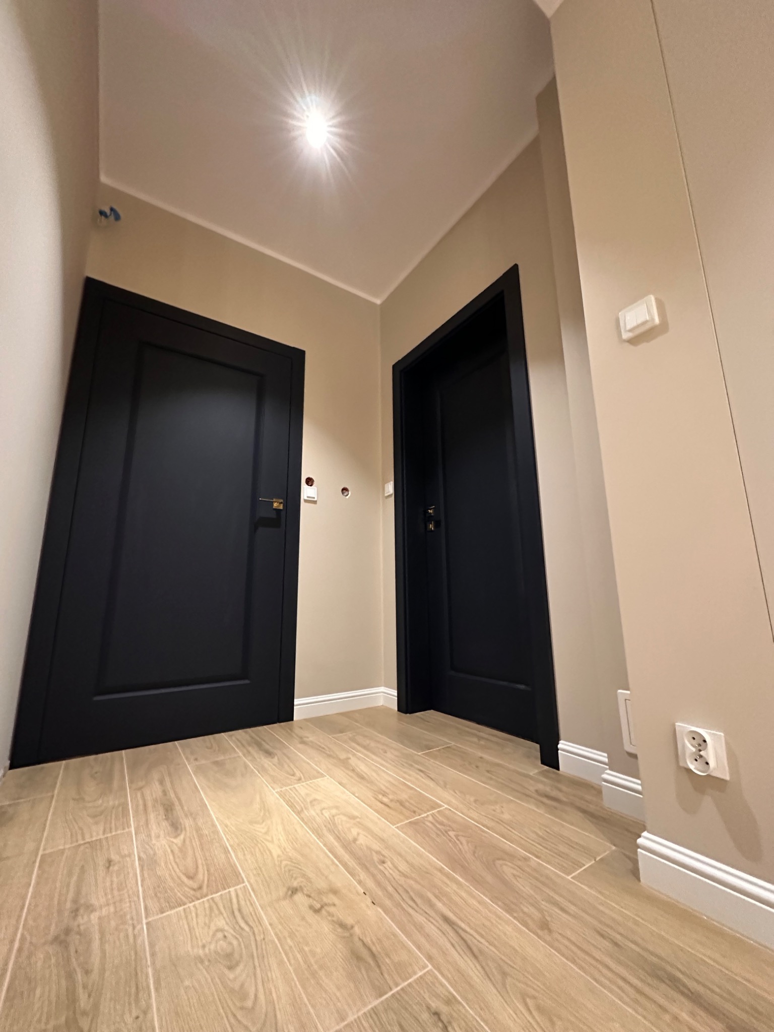 Korytarz po remoncie z dwiema czarnymi drzwiami, podłoga wyłożona płytkami imitującymi drewno, ściany w odcieniach beżu, widoczne gniazdka elektryczne i włącznik światła.