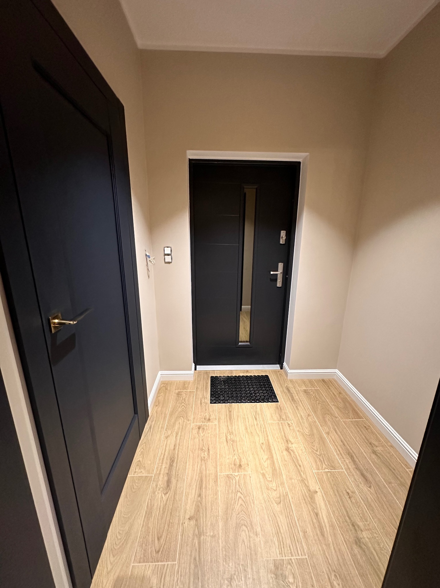 Wnętrze przedpokoju z drewnopodobną podłogą, czarnymi drzwiami wejściowymi z szybką i matą podłogową oraz drugimi czarnymi drzwiami z złotą klamką po lewej stronie.