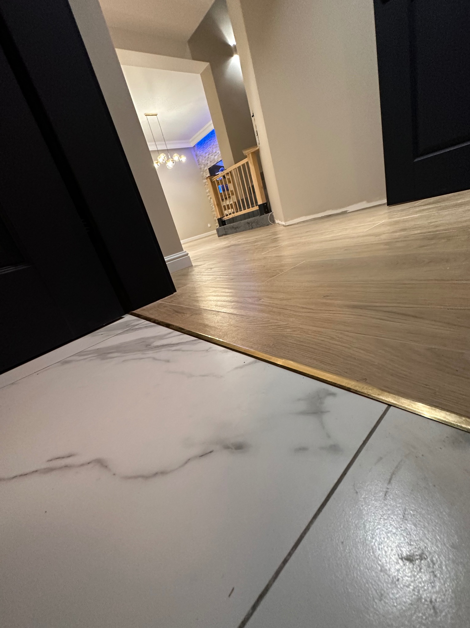 Połączenie podłogi z płytek imitujących marmur z panelami podłogowymi, wykończone listwą progową w kolorze złotym, widok na przedpokój z oświetleniem.