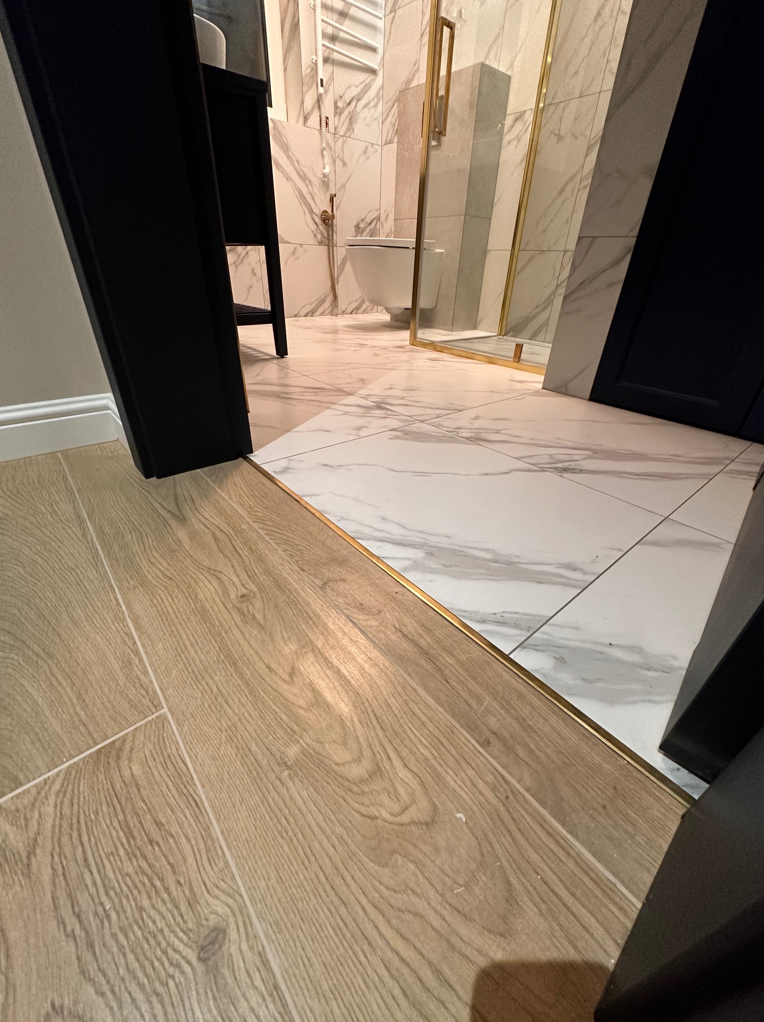 Połączenie podłogi drewnianej z marmurową w wyremontowanej łazience z kabiną prysznicową ze złotymi elementami, widoczne przejście z listwą wykończeniową.