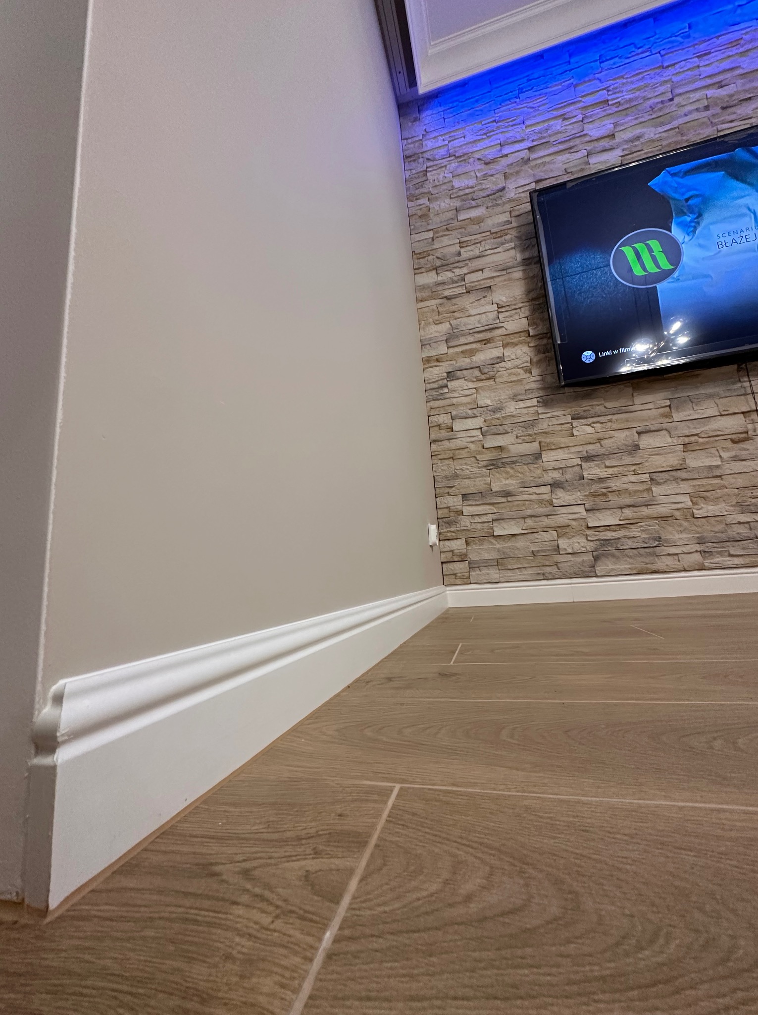 Ściana w jasnym odcieniu z białą listwą przypodłogową w Gdyni, wykończona panelami imitującymi kamień, na której zawieszony jest telewizor z podświetleniem LED.