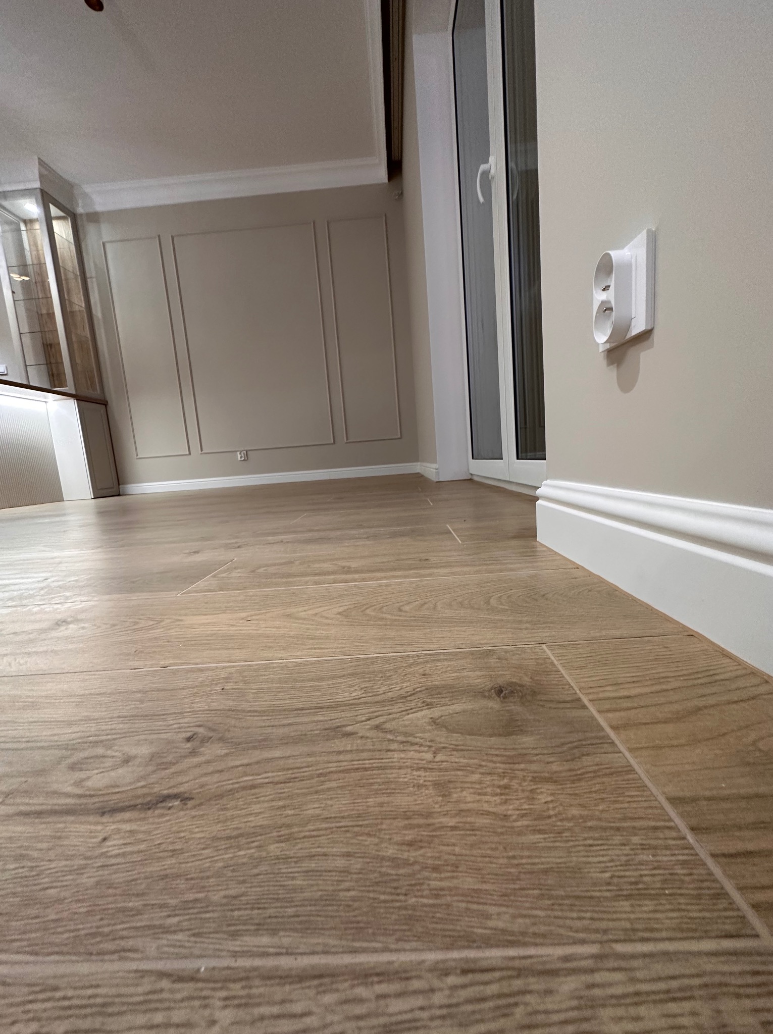 Nowoczesne wnętrze po remoncie: drewnopodobna podłoga, listwy przypodłogowe, gładkie ściany w odcieniach beżu z dekoracyjnymi panelami i białe gniazdko elektryczne z osłoną.