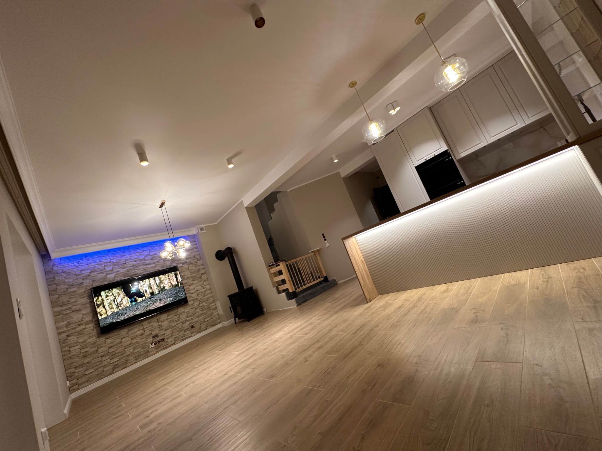 Nowoczesne wnętrze salonu z aneksem kuchennym: drewniana podłoga, ściana z kamienia dekoracyjnego podświetlona niebieskim światłem, telewizor, czarny piec wolnostojący, biała zabudowa kuchenna...