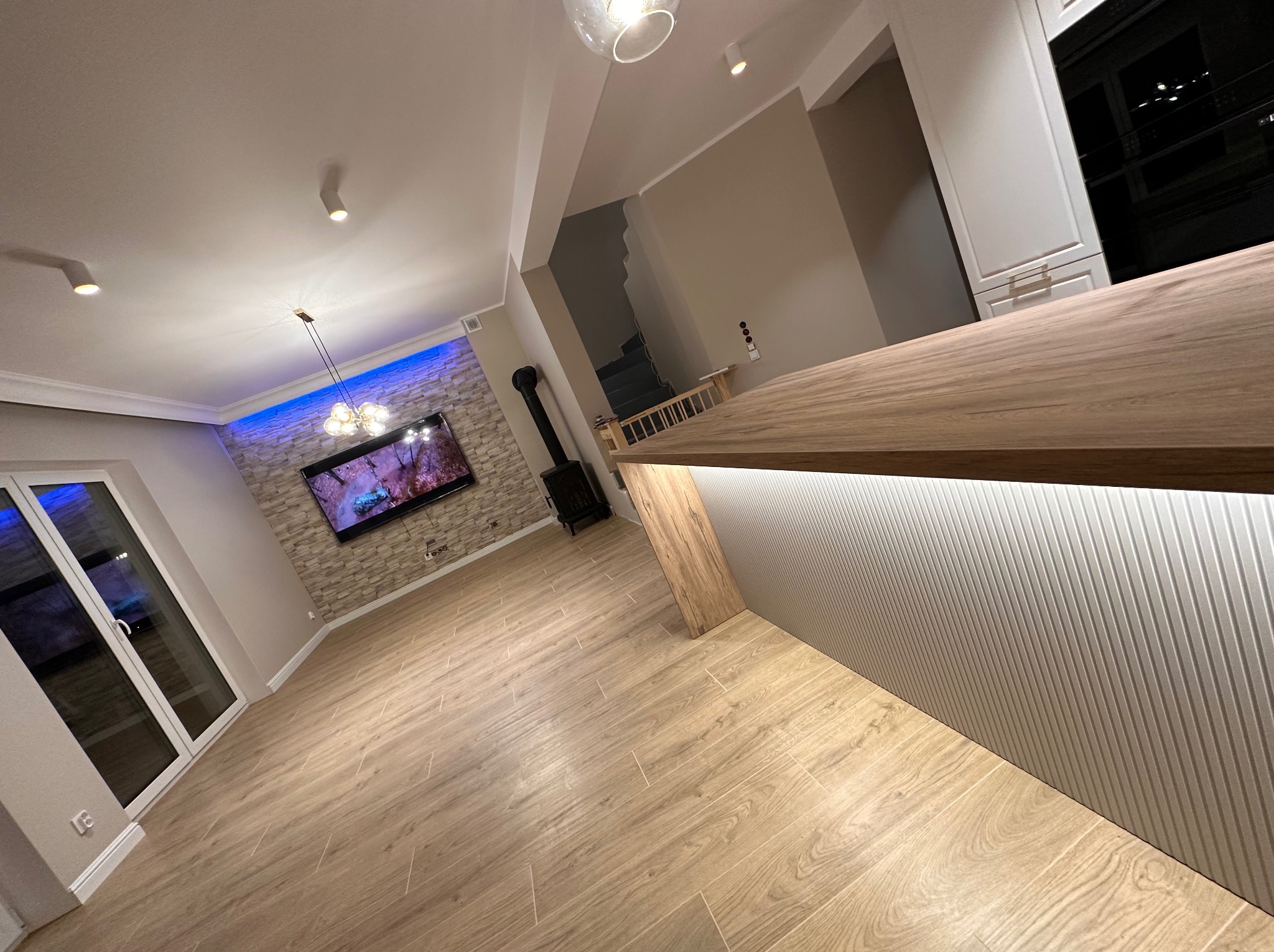 Przestronne wnętrze po remoncie z drewnianą podłogą, ścianą z kamienia dekoracyjnego podświetloną niebieskim światłem, nowoczesnym kominkiem i wyspą kuchenną z oświetleniem LED.