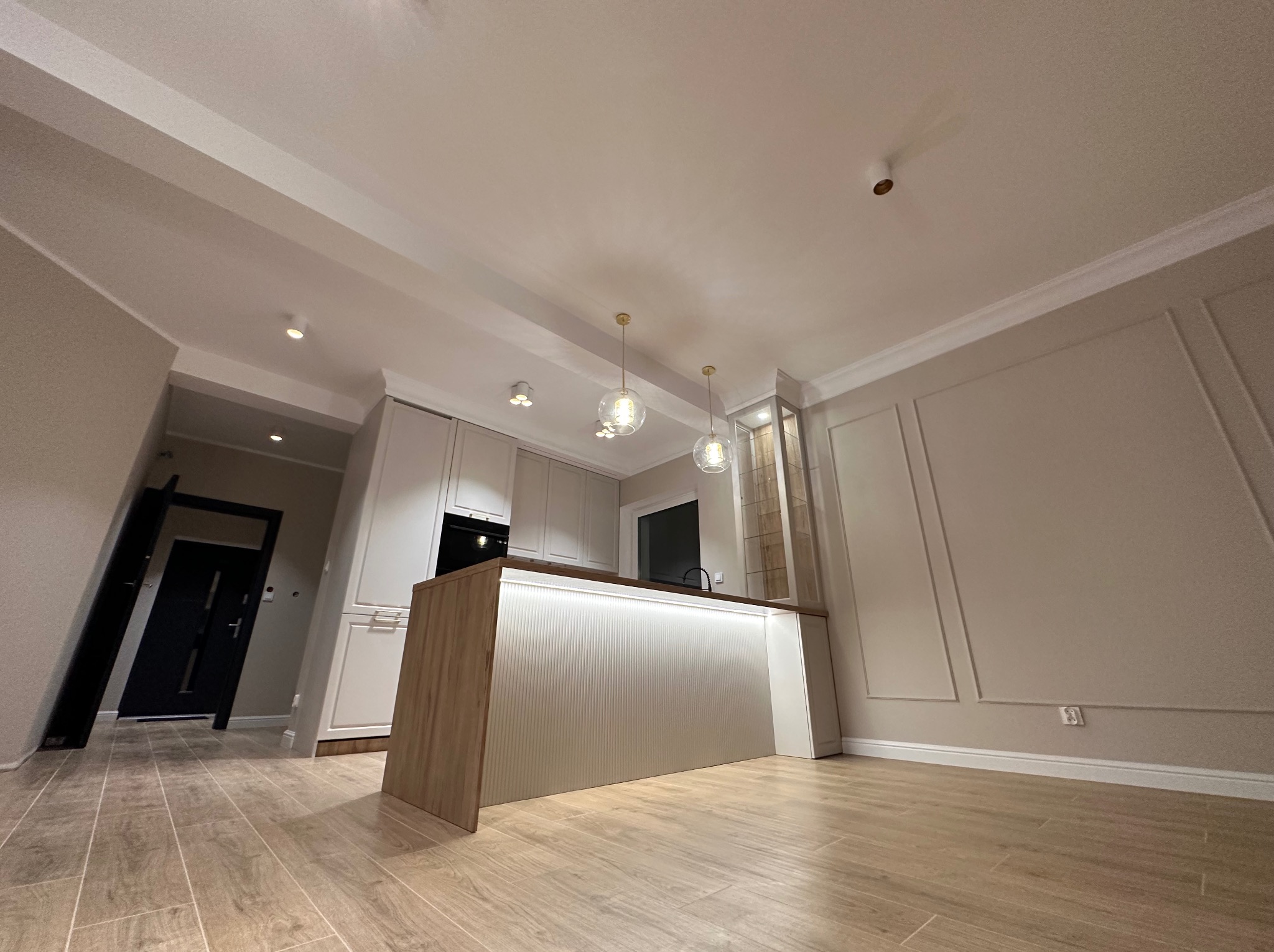 Nowoczesna kuchnia z wyspą oświetloną listwą LED, wiszącymi lampami kulami i dekoracyjnymi panelami ściennymi.