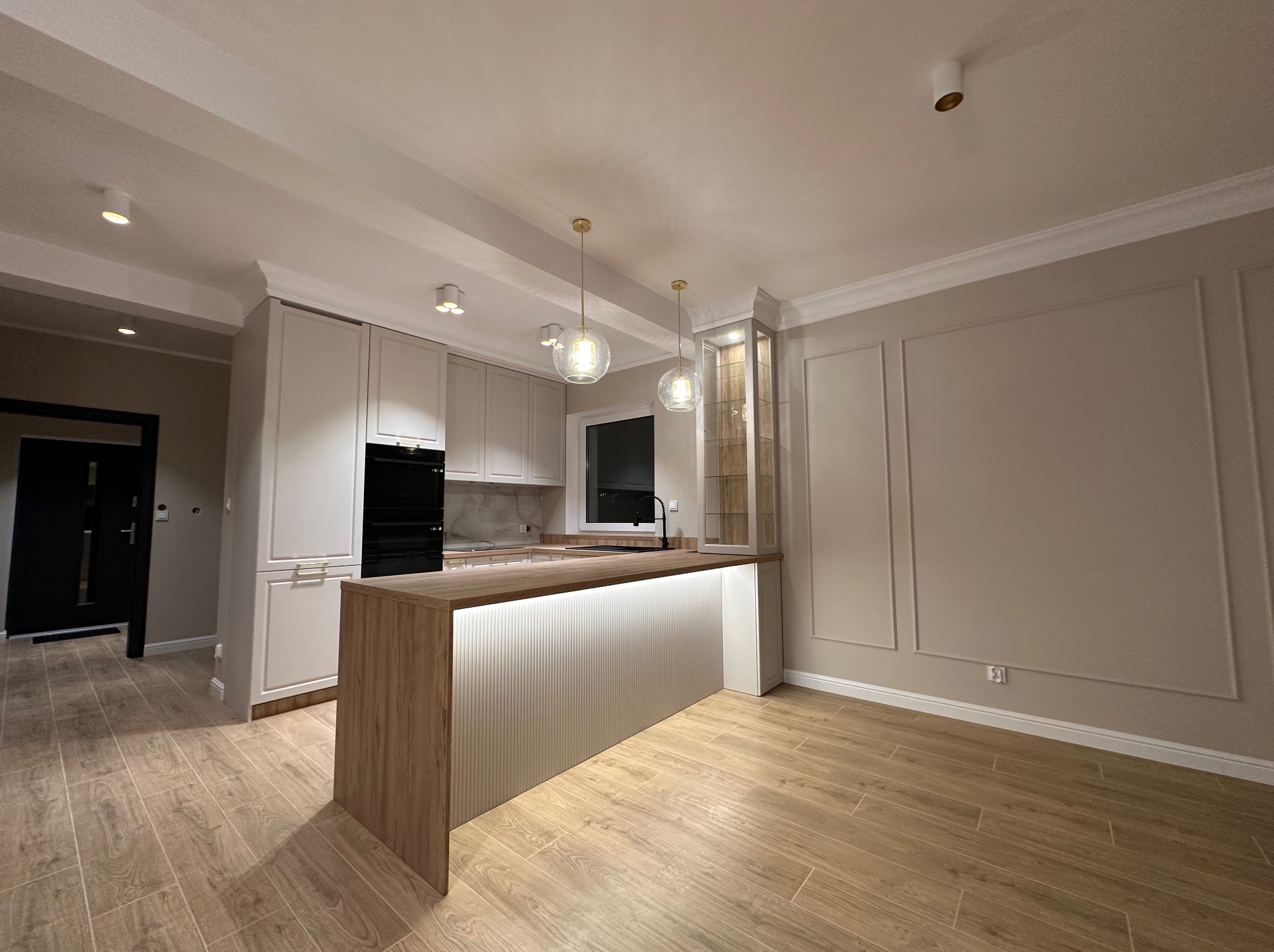 Nowoczesna kuchnia z wyspą, oświetleniem LED pod blatem i dekoracyjnymi listwami ściennymi, wykończona w jasnych barwach.