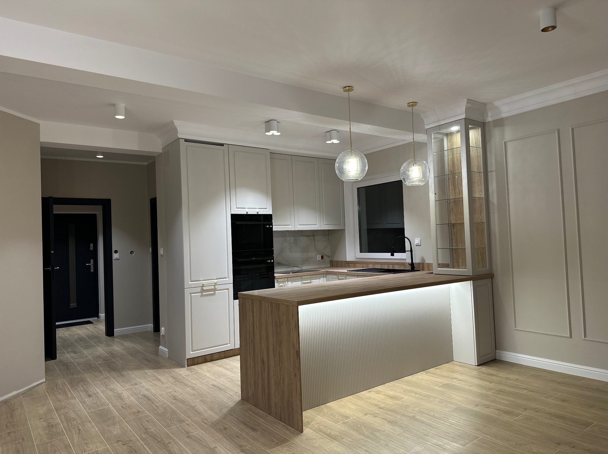 Nowoczesna kuchnia z wyspą, białe szafki z frezowanymi frontami, drewniany blat z podświetleniem LED, wiszące lampy kule, płytki imitujące drewno na podłodze, dekoracyjne listwy na ścianach.