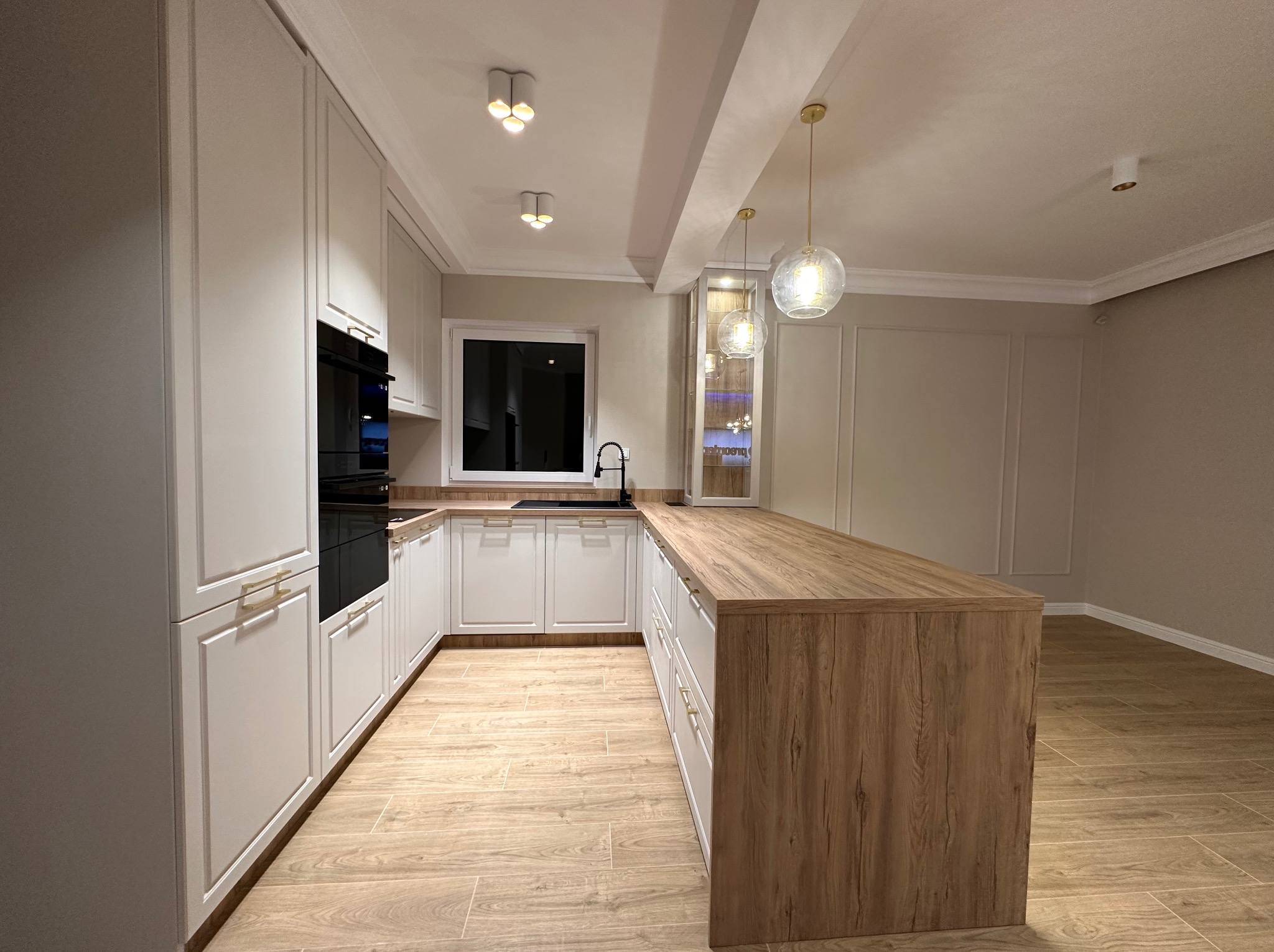 Nowoczesna kuchnia z białymi szafkami, drewnianym blatem wyspy i złotymi akcentami, oświetlona wiszącymi lampami w kształcie kul.